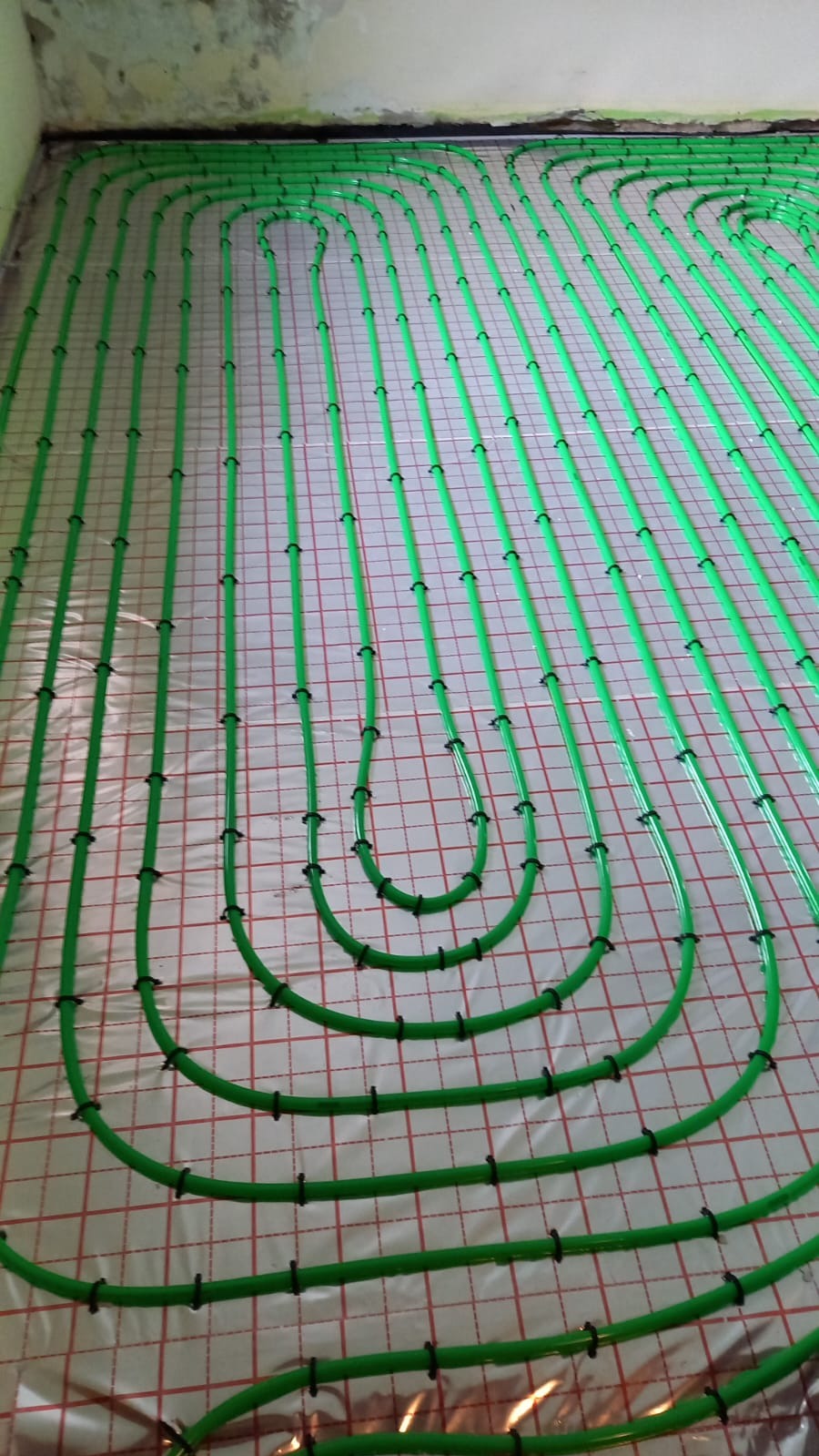 Ułożone, zielone rury ogrzewania podłogowego na folii z czerwoną siatką, przymocowane czarnymi uchwytami. Widok z góry, instalacja przed wylaniem posadzki.