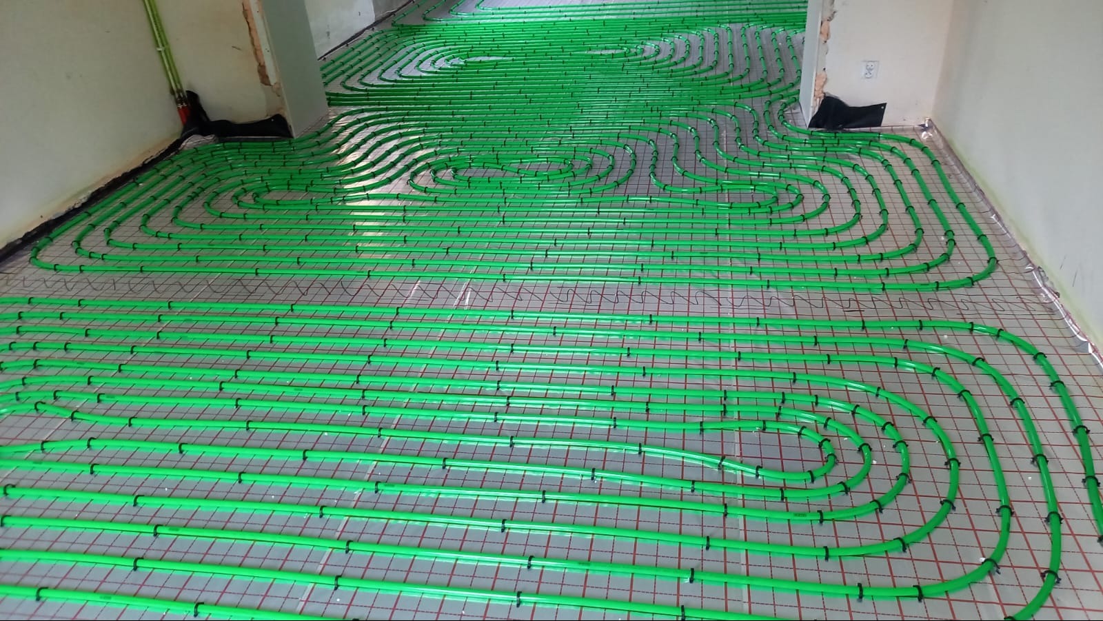 Zielone rury ogrzewania podłogowego ułożone na siatce, gotowe do zalania posadzką, widoczne fragmenty ścian.