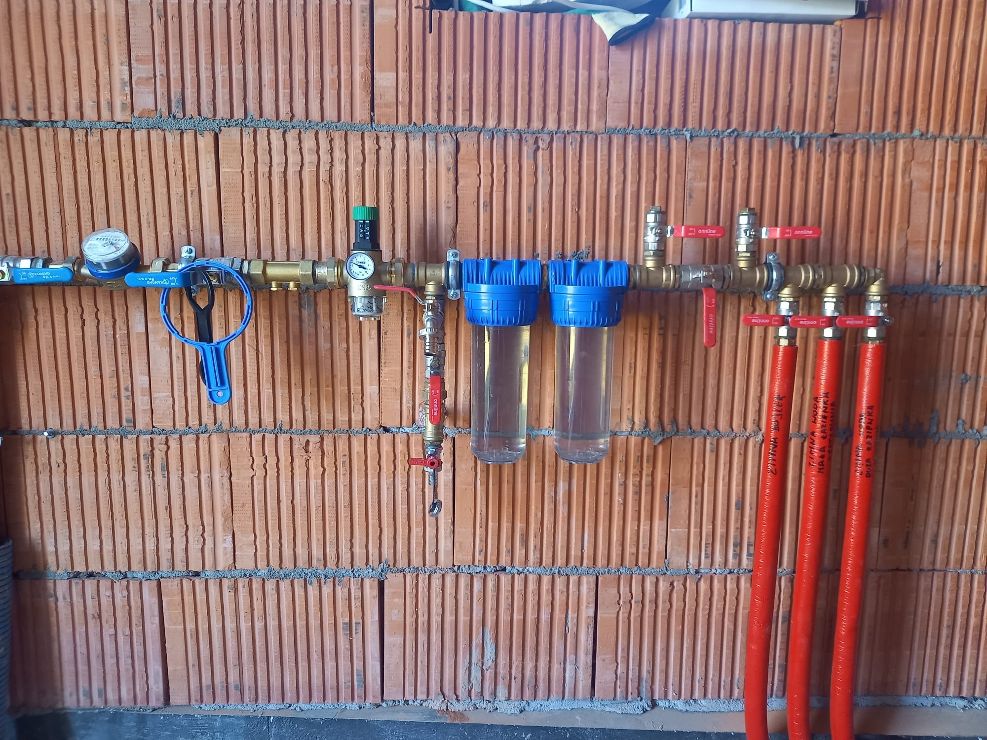 Instalacja wodna z filtrami i zaworami na tle ściany z cegły dziurawki. Widoczne czerwone rury z opisami, manometr i niebieski klucz do filtrów.