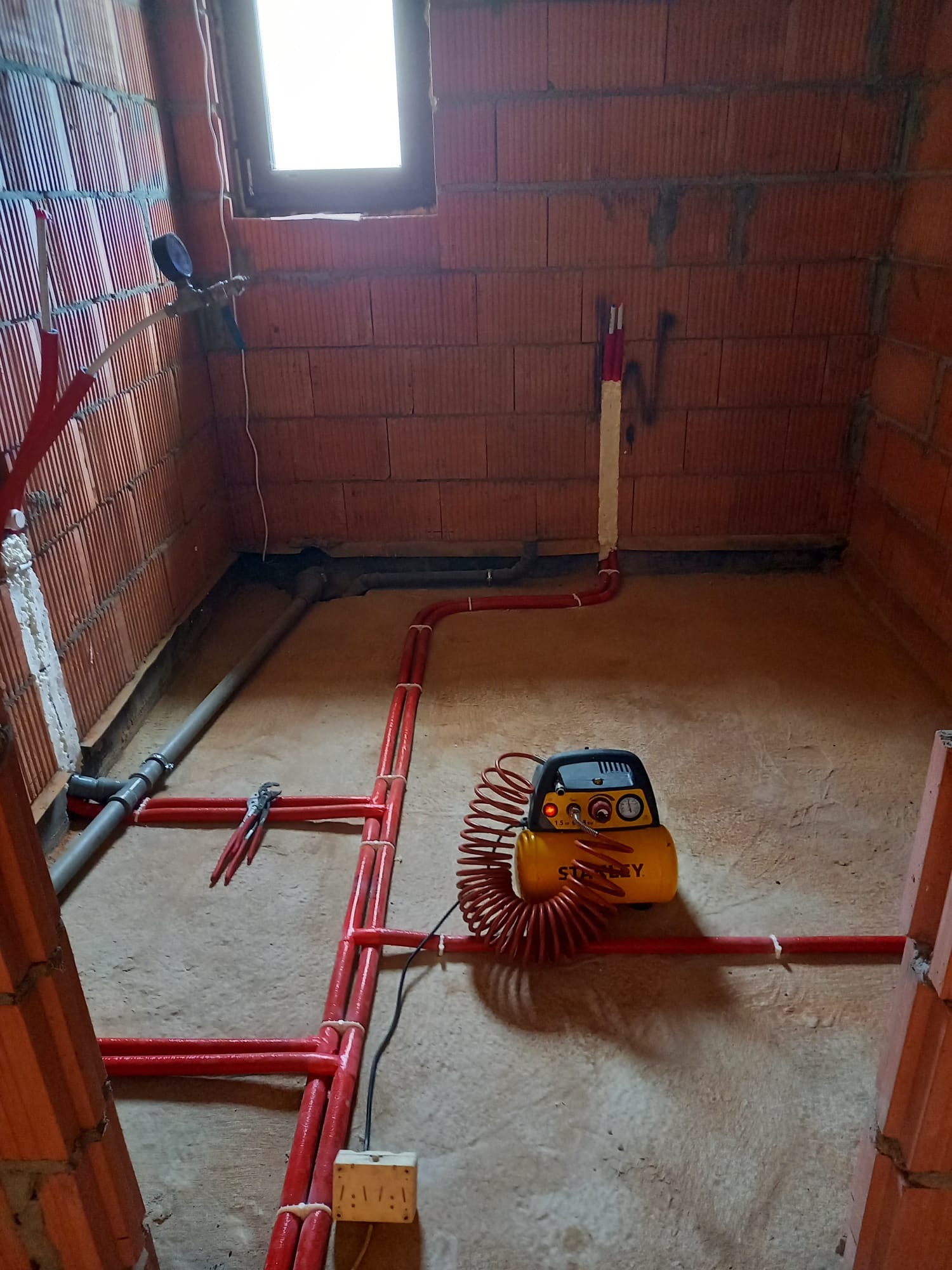 Czerwone rury instalacji ogrzewania podłogowego w surowym wnętrzu z ceglanymi ścianami. Kompresor Stanley i szczypce obok. Widok z góry na podłogę.