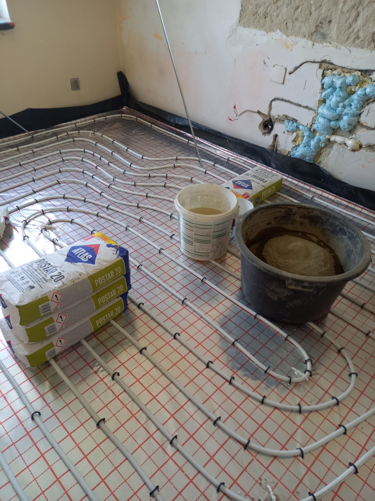 Ułożone rury ogrzewania podłogowego na siatce, obok worki z zaprawą Atlas Postar 20 i wiadro z rozrobioną zaprawą. Ściana z instalacją zabezpieczona pianą.