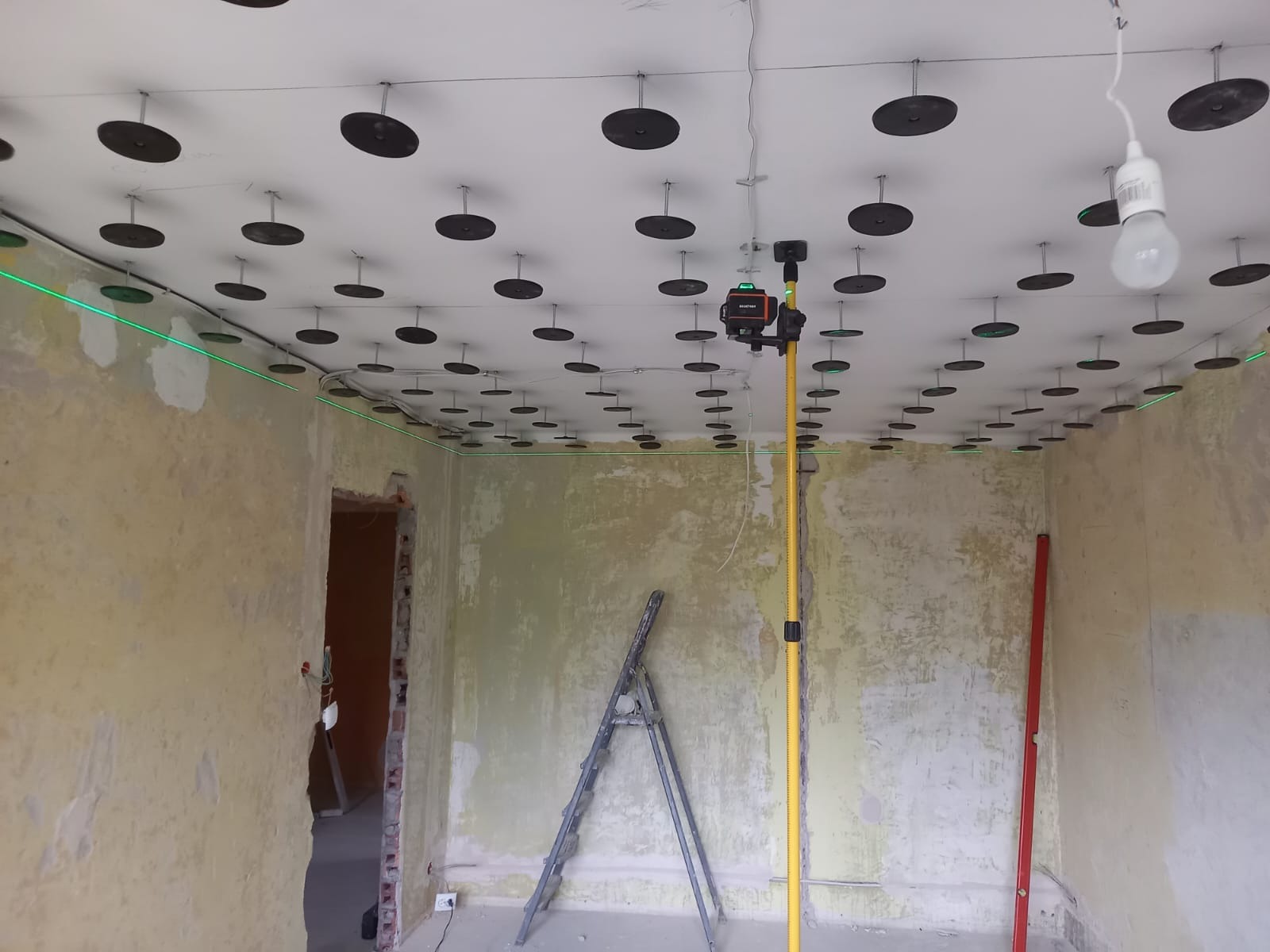 Montaż sufitu podwieszanego z użyciem lasera krzyżowego. Widoczne profile i elementy mocujące na suficie. Ściany w stanie surowym, drabina oparta o ścianę.