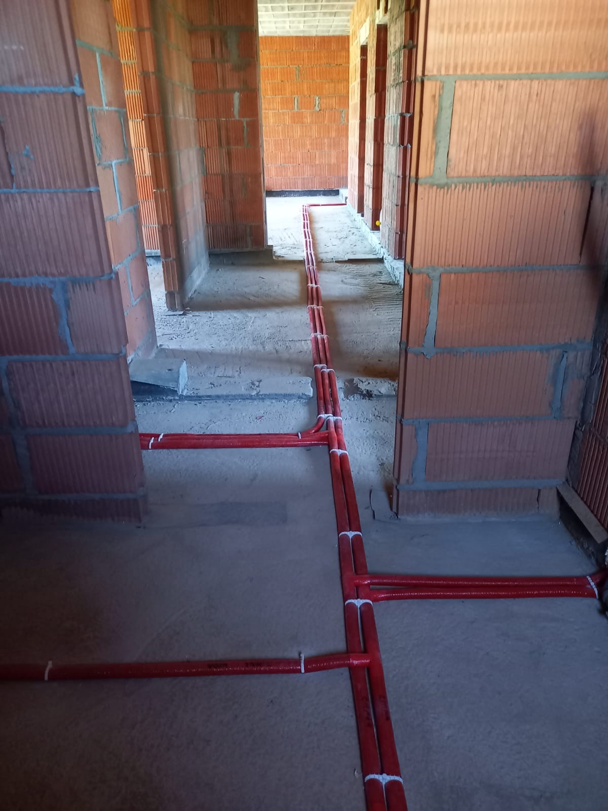 Czerwone rury ogrzewania podłogowego, ułożone na szarym podłożu w surowym wnętrzu budynku z czerwonej cegły. Widok z góry na instalację przed wylaniem posadzki.
