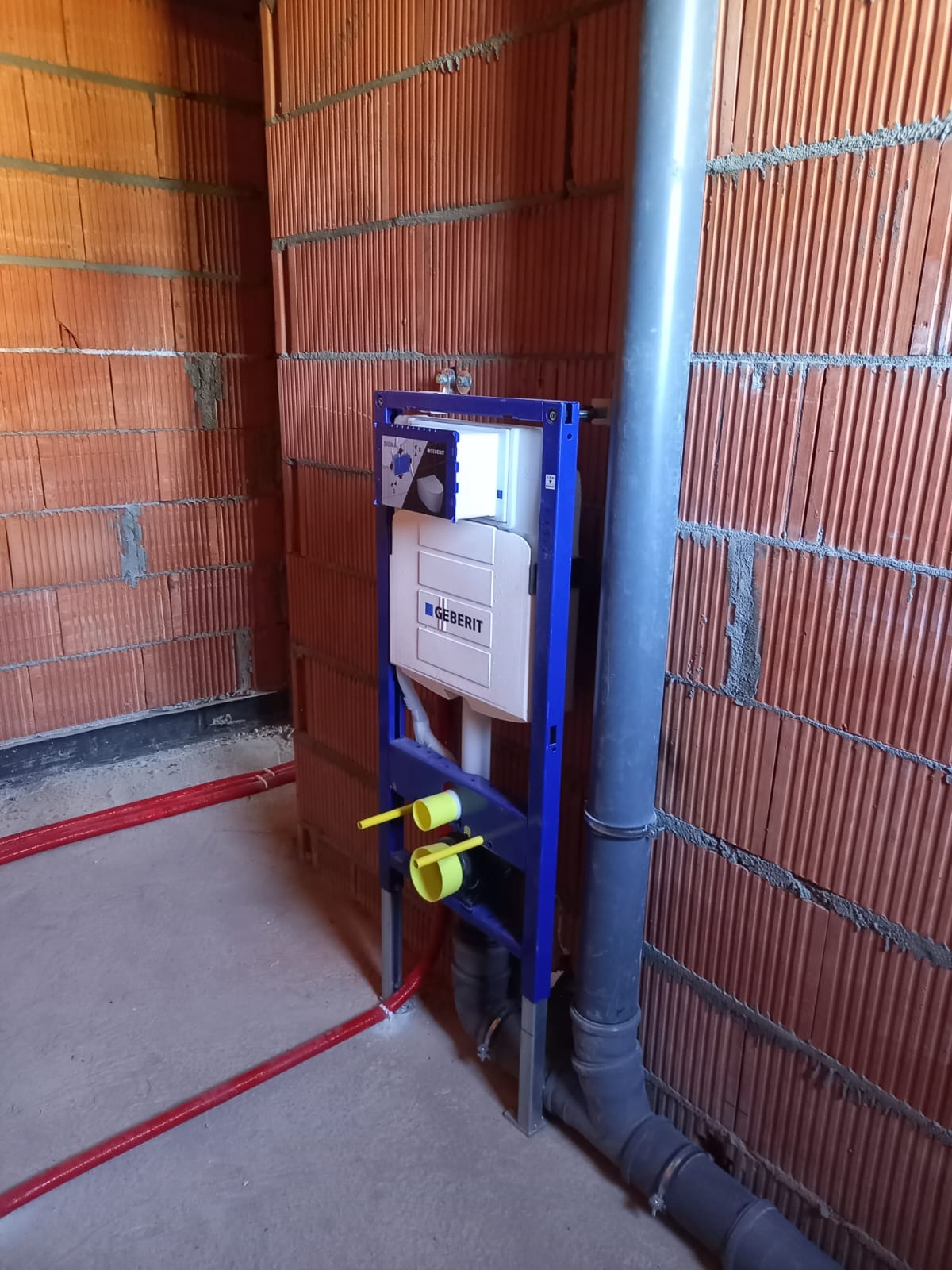Stelaż podtynkowy Geberit Duofix zamontowany przy czerwonej ścianie z pustaków. Widoczne rury kanalizacyjne i dopływowe. Instalacja w trakcie montażu.