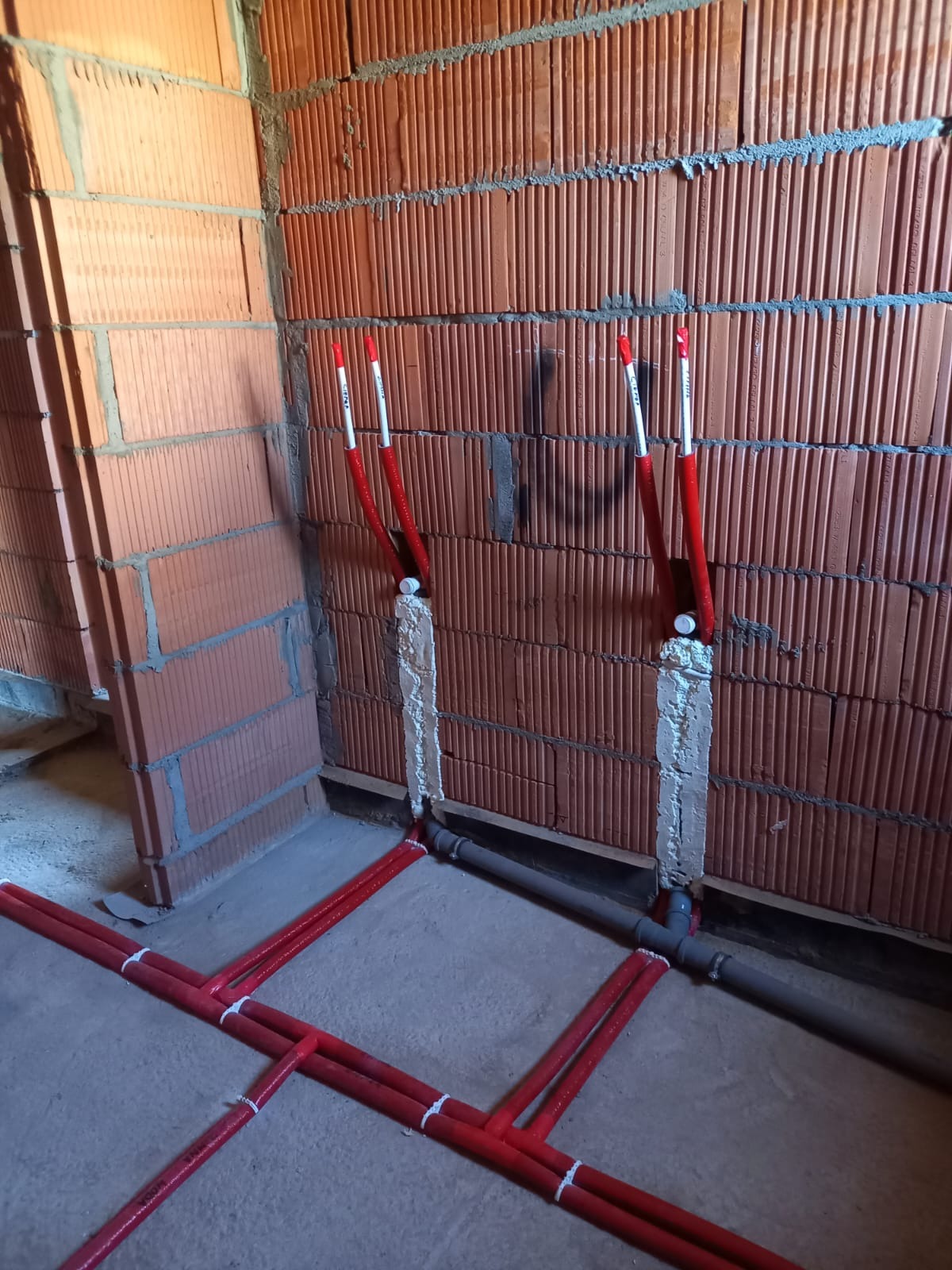 Czerwone rury instalacji wodnej zamocowane do ściany z cegły, zabezpieczone pianką montażową. Szare rury kanalizacyjne wzdłuż podłogi.