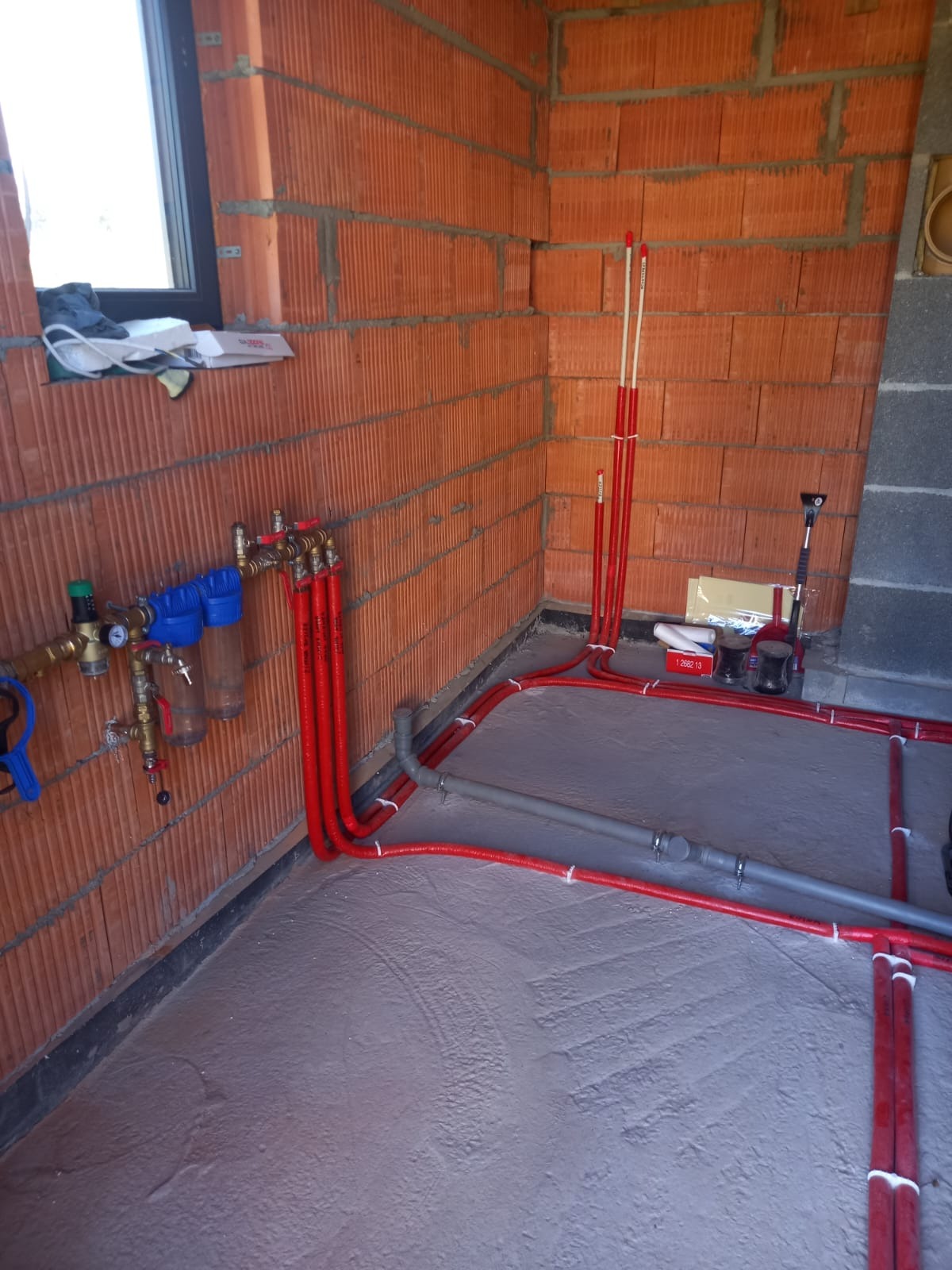 Czerwone rury instalacji grzewczej ułożone na szarym podkładzie podłogowym w surowym wnętrzu z ceglanymi ścianami, widoczne filtry i zawory.