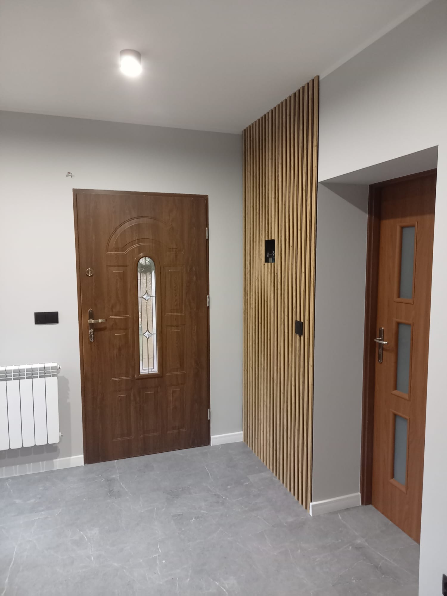 Szare wnętrze: marmurowe płytki, drewniana lamelowa ściana, brązowe drzwi wejściowe i wewnętrzne, biały grzejnik. Minimalistyczny design.