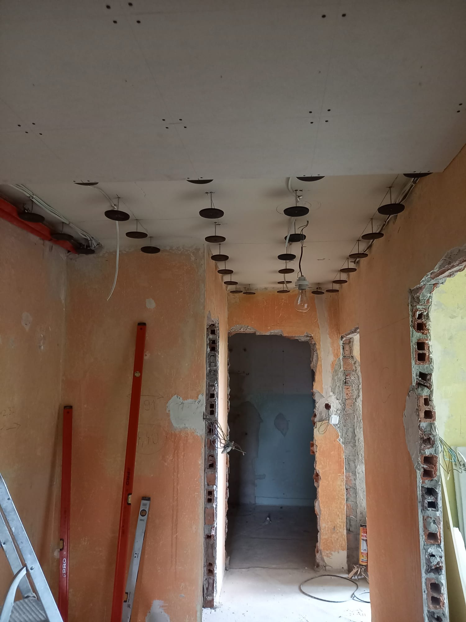 Remont sufitu podwieszanego z widocznymi elementami mocującymi, instalacjami i ścianami w stanie surowym. W tle widoczne przejście do kolejnego pomieszczenia.