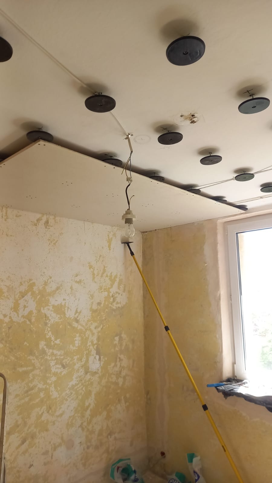 Montaż podwieszanego sufitu z widocznymi elementami mocującymi i prowizorycznym oświetleniem w pomieszczeniu w trakcie remontu. Ściany częściowo przygotowane do malowania.