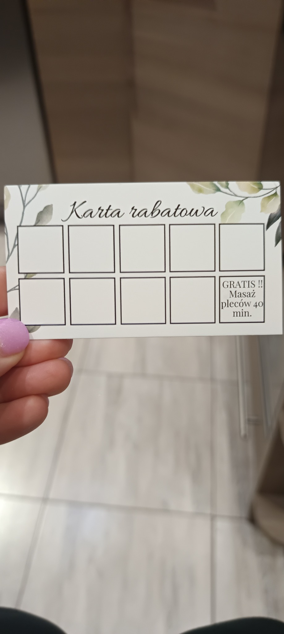 Karta rabatowa z motywem roślinnym, oferująca darmowy masaż pleców 40 min po zebraniu pieczątek. Trzymana w dłoni z pomalowanym paznokciem.
