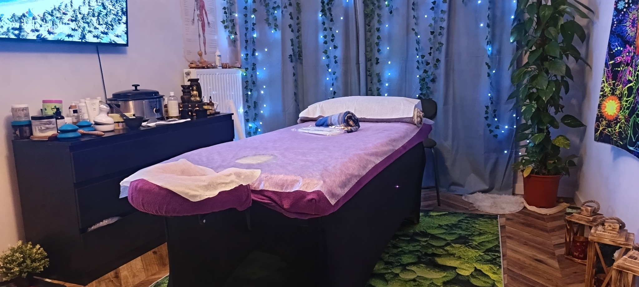 Przytulne wnętrze gabinetu z łóżkiem do zabiegów, dekoracjami roślinnymi i nastrojowym oświetleniem, tworzące relaksującą atmosferę. Widoczne akcesoria do zabiegów.