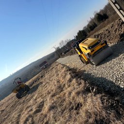 Pat-Trans - Żółty walec drogowy ubija kamienistą nawierzchnię na zboczu, w tle widać wywrotkę i drzewa. Błękitne niebo i słup energetyczny dopełniają krajobraz.