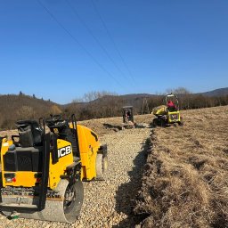 Pat-Trans - Żółty walec JCB ubija kamienistą drogę na polu, w tle pracuje mini-wywrotka i koparka, błękitne niebo, linia wysokiego napięcia. Utwardzanie drogi w terenie.