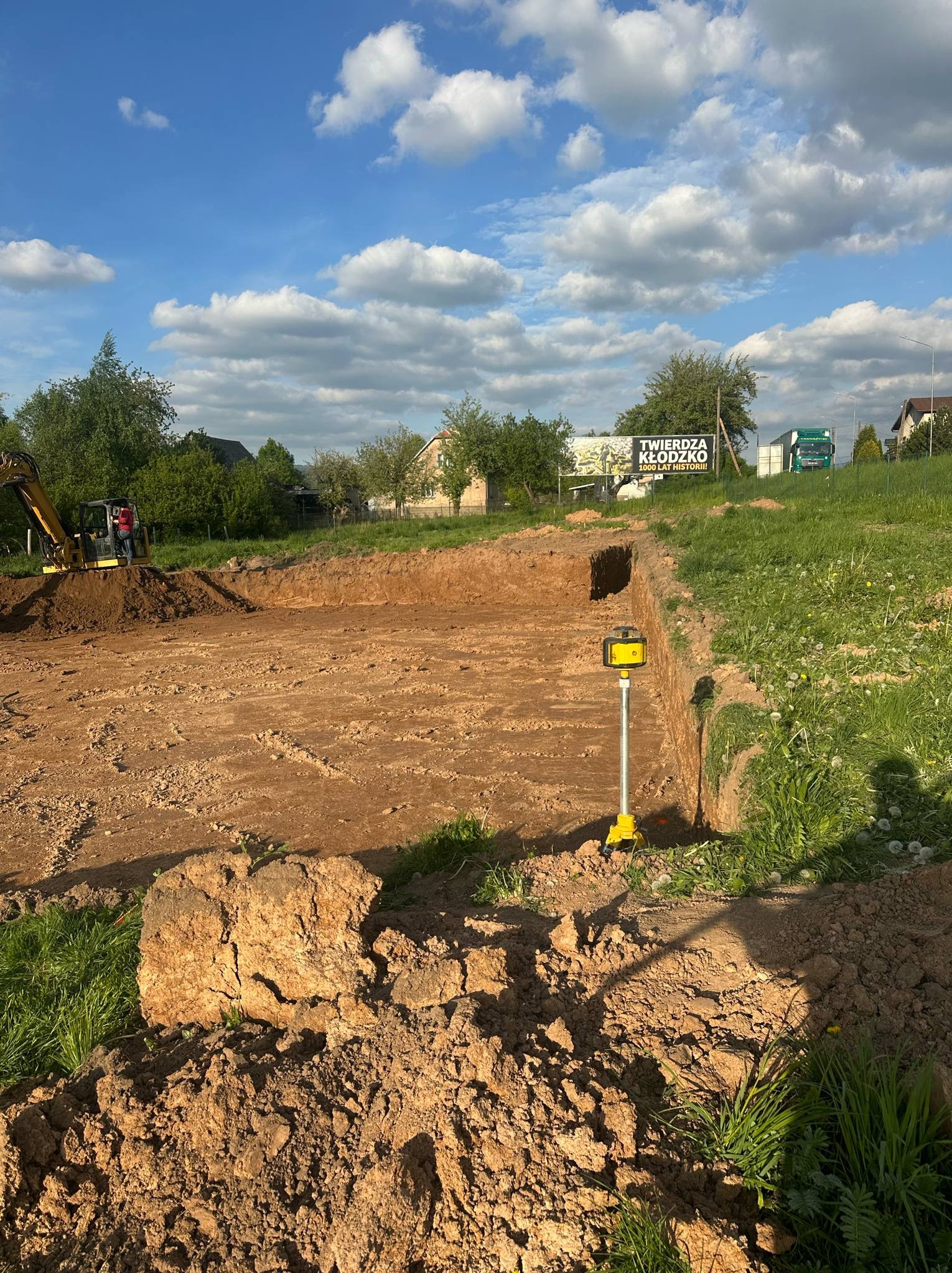 Żółta niwelatorka laserowa w wykopie pod fundamenty, w tle koparka i tablica reklamowa 'Twierdza Kłodzko'. Błękitne niebo z chmurami.