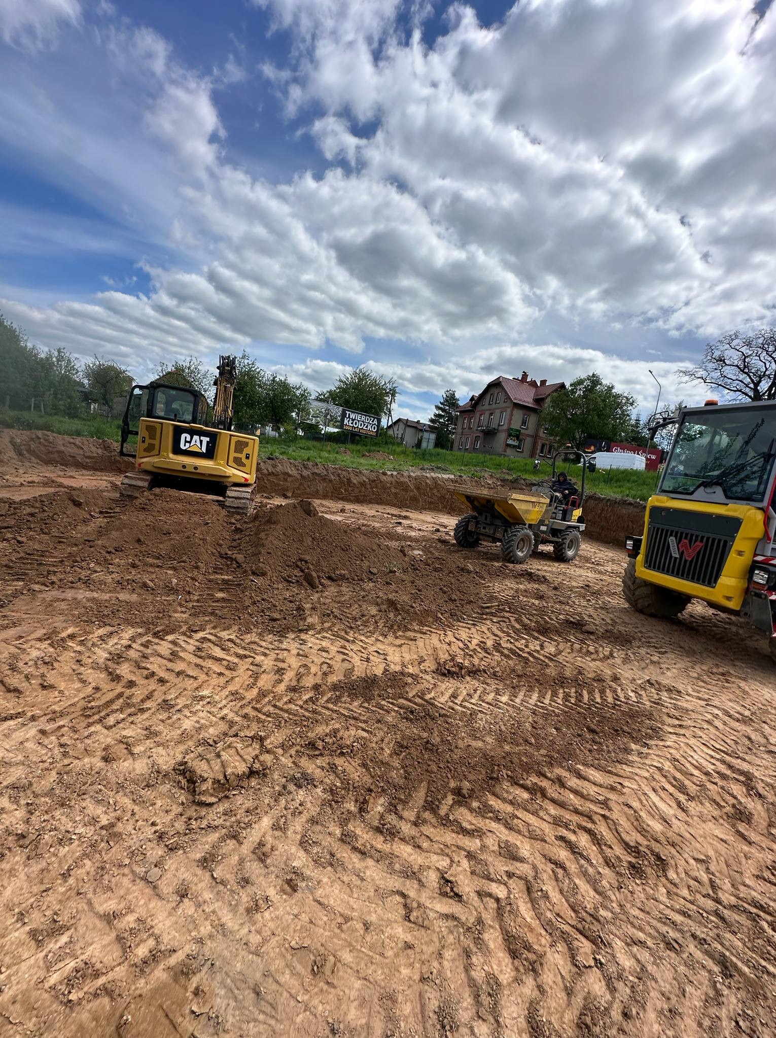 Wykop pod budowę w Szczytnej z widocznymi maszynami budowlanymi: koparka CAT, wywrotka i walec. Ślady opon na ziemi. Błękitne niebo z chmurami w tle.