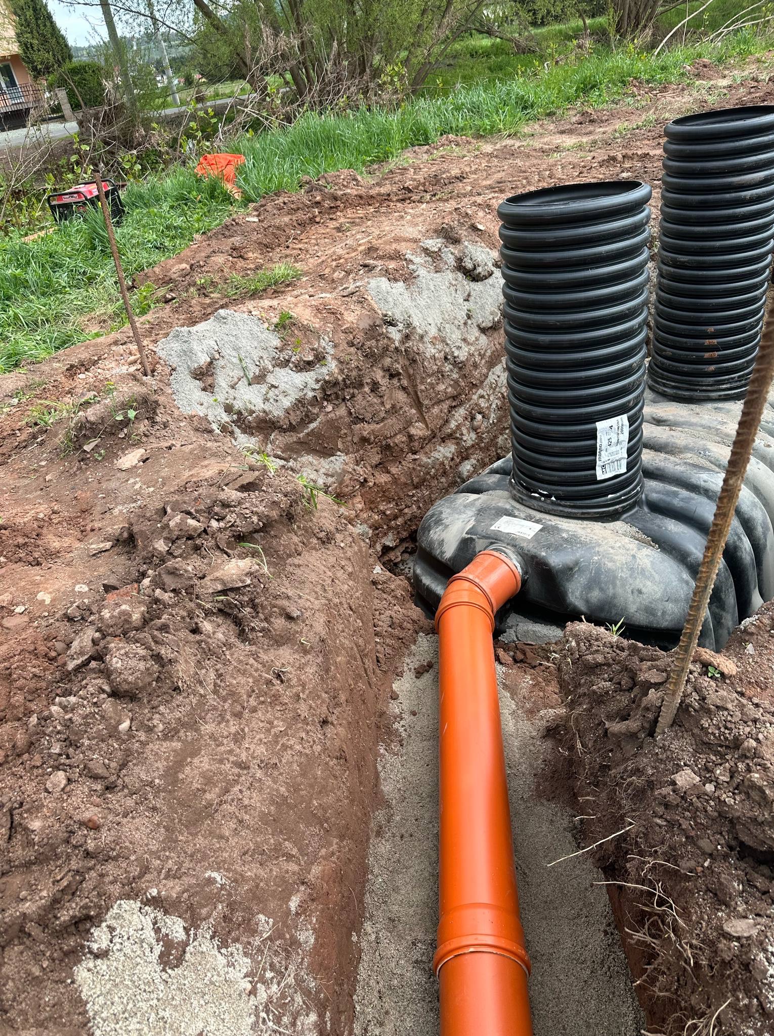 Montaż szamba: wykop z piaskowym podsypem, podłączona rura kanalizacyjna, zbiornik z dwoma kominami wentylacyjnymi. Widoczny generator prądu w tle.