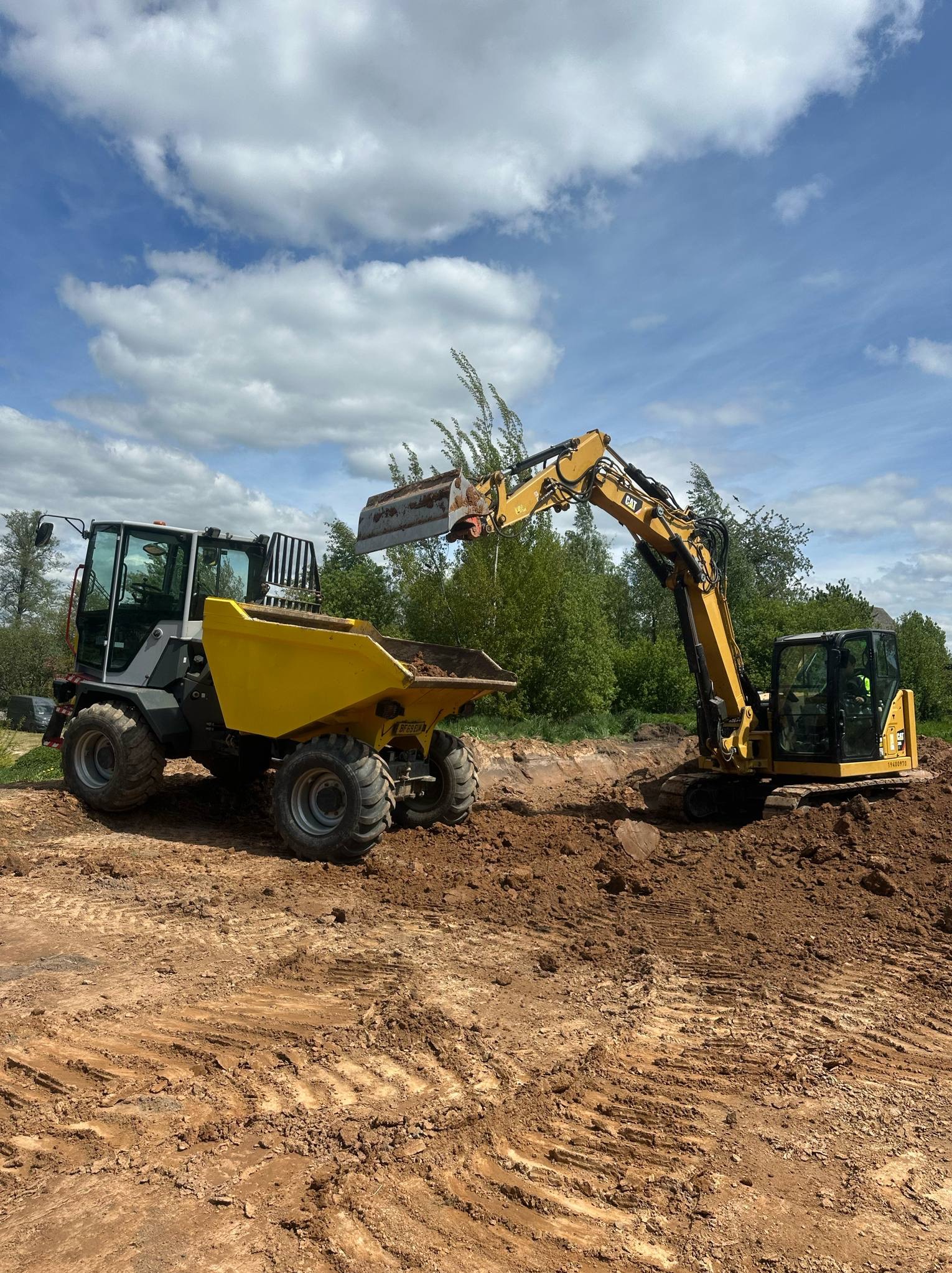 Żółty wywrotka i koparka CAT pracują na placu budowy w słoneczny dzień. Koparka nabiera ziemię i przekazuje ją do wywrotki. Widoczne ślady opon na ziemi.