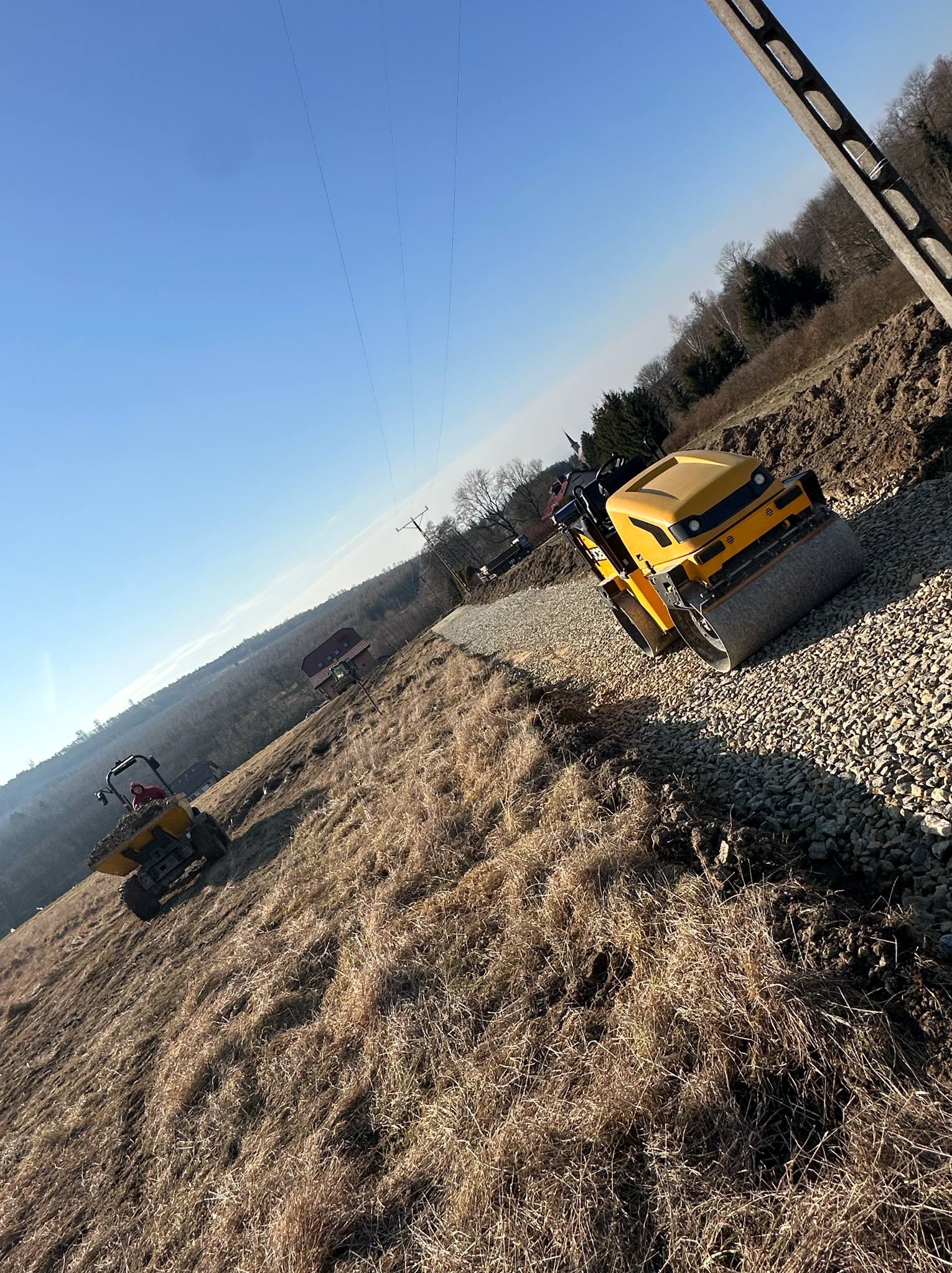 Żółty walec drogowy ubija kamienistą nawierzchnię na zboczu, w tle widać wywrotkę i drzewa. Błękitne niebo i słup energetyczny dopełniają krajobraz.