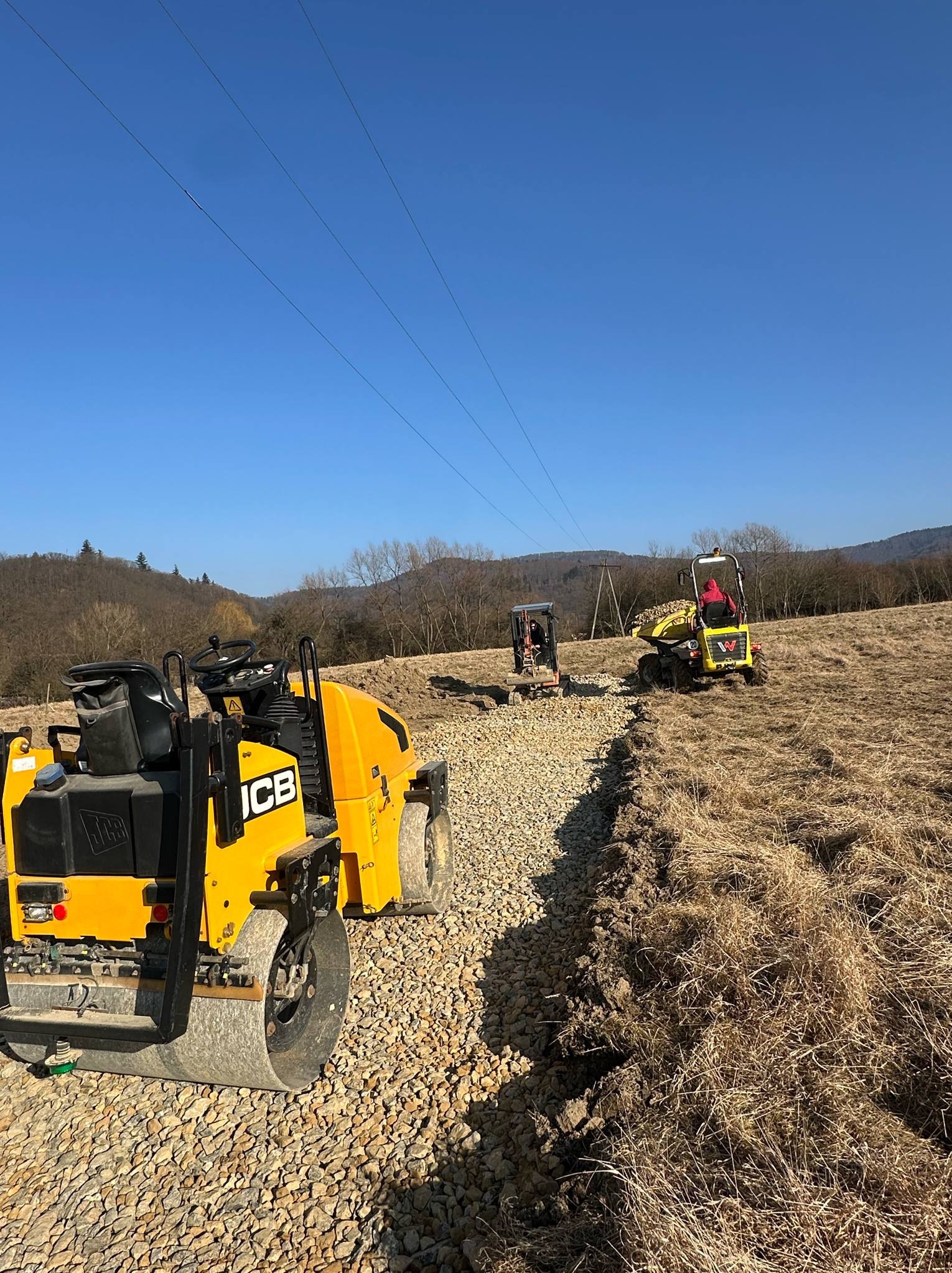 Żółty walec JCB ubija kamienistą drogę na polu, w tle pracuje mini-wywrotka i koparka, błękitne niebo, linia wysokiego napięcia. Utwardzanie drogi w terenie.