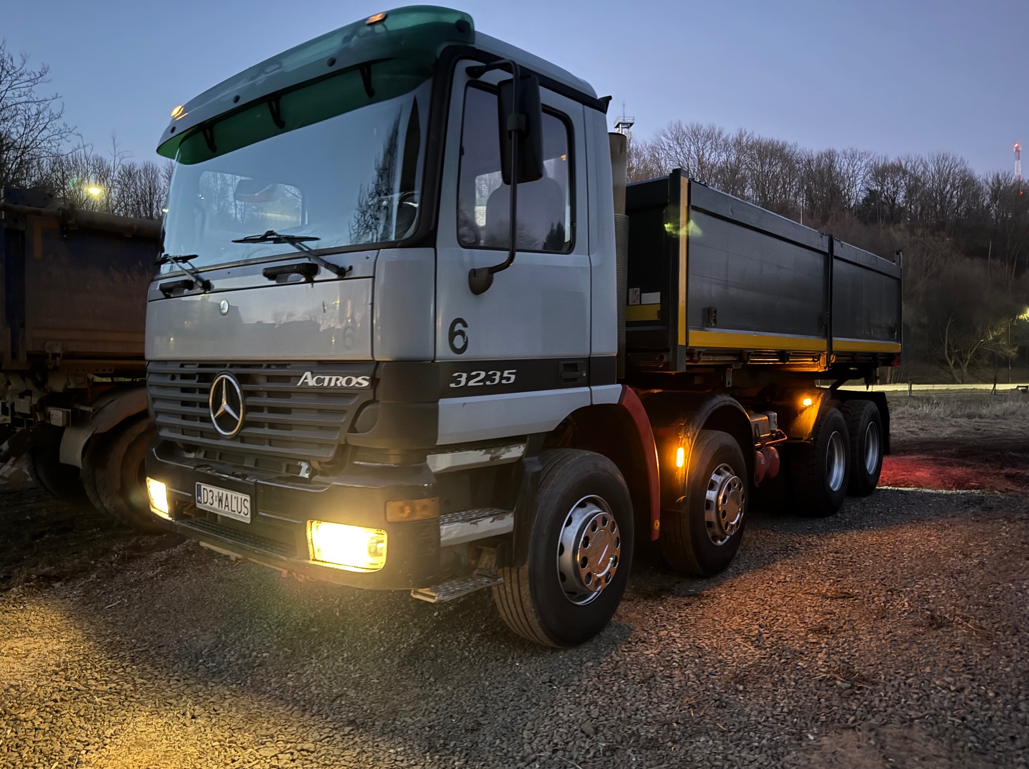 Szary wywrotka Mercedes Actros 3235 z włączonymi światłami, zaparkowany na żwirowym placu. W tle drzewa i elementy infrastruktury.