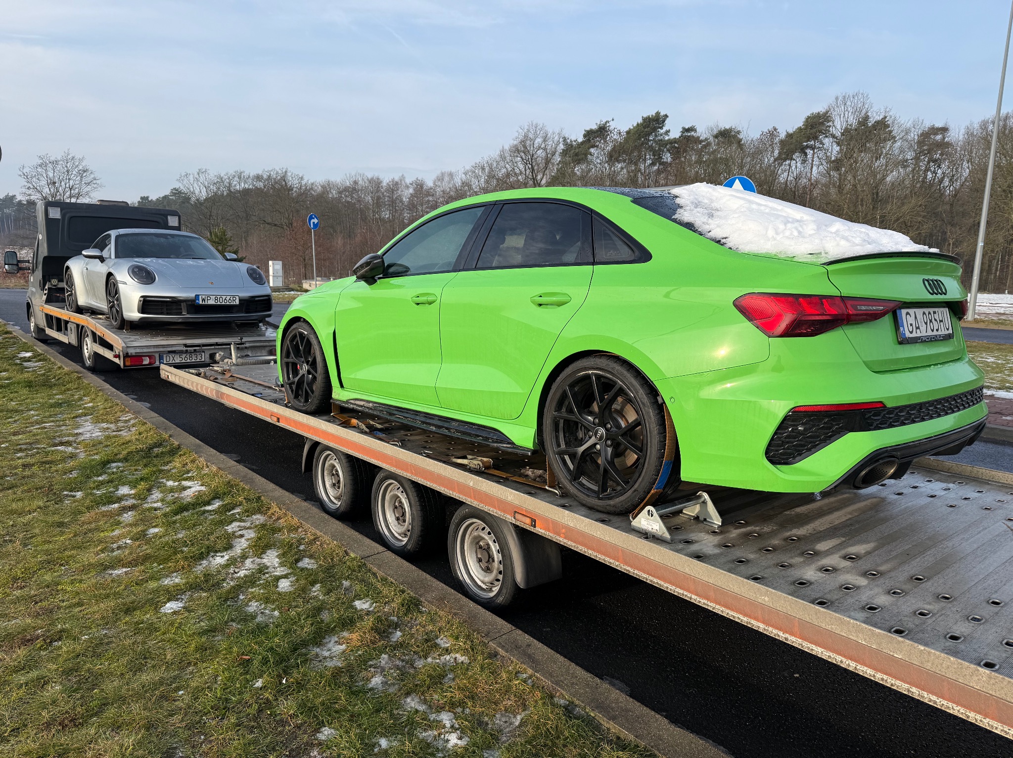 Laweta przewozi zielone Audi RS3 z zaspą śniegu na dachu i srebrne Porsche. Widoczne tablice rejestracyjne. Transport aut na lawecie w zimowych warunkach.