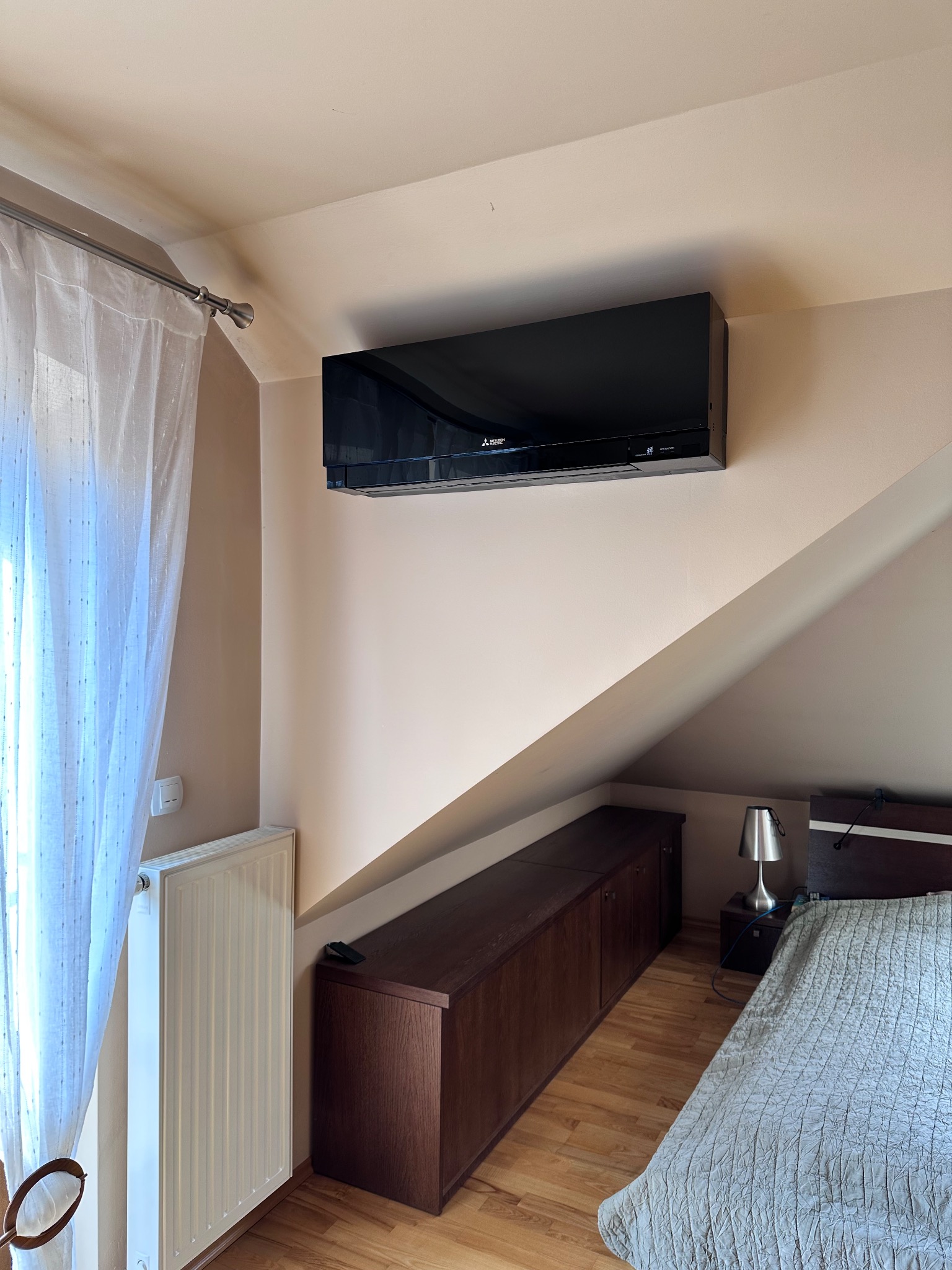Czarny klimatyzator Mitsubishi Electric zamontowany na ścianie w sypialni na poddaszu, widoczny fragment łóżka i okno z białą zasłoną.