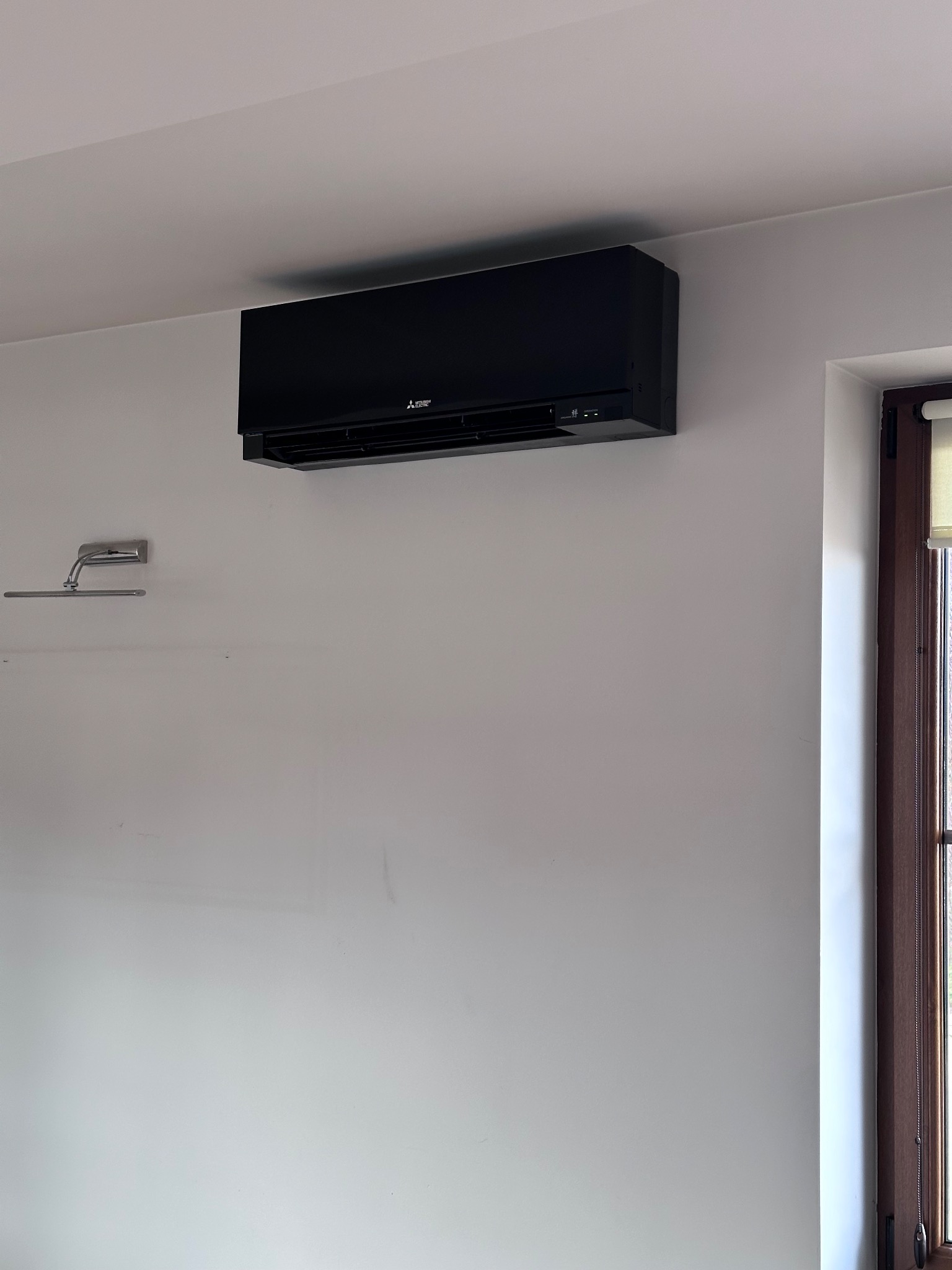 Czarny klimatyzator Mitsubishi zamontowany na białej ścianie, obok okna z drewnianą ramą i roletą. Widoczna lampka nad szafką.