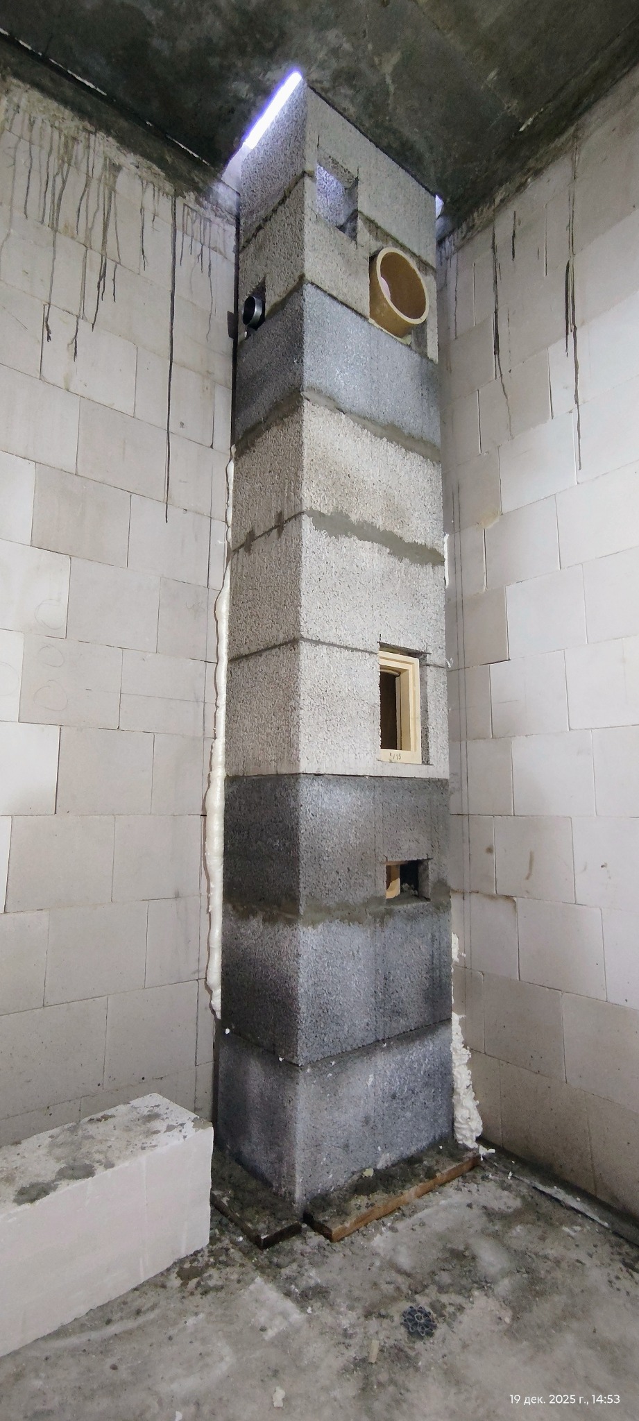 Wznoszony komin z szarych bloczków betonowych w surowym wnętrzu. Widoczne otwory wentylacyjne i rura. Ściany z białych bloczków, szczelina wypełniona pianką montażową.