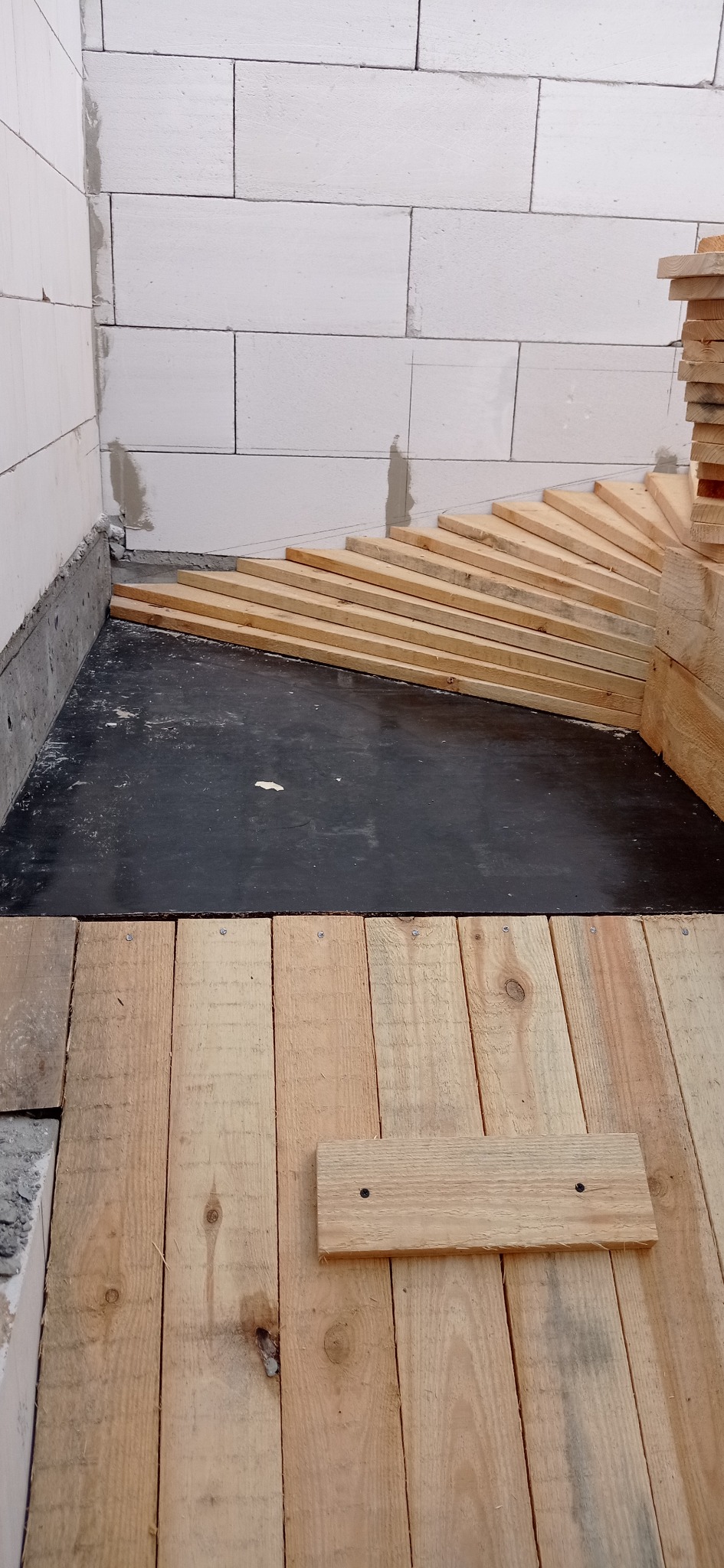 Budowa podłogi w domu z bloczków: izolacja z czarnej folii, ułożone deski podłogowe, widoczne ściany z betonu komórkowego. Etap prac wykończeniowych.