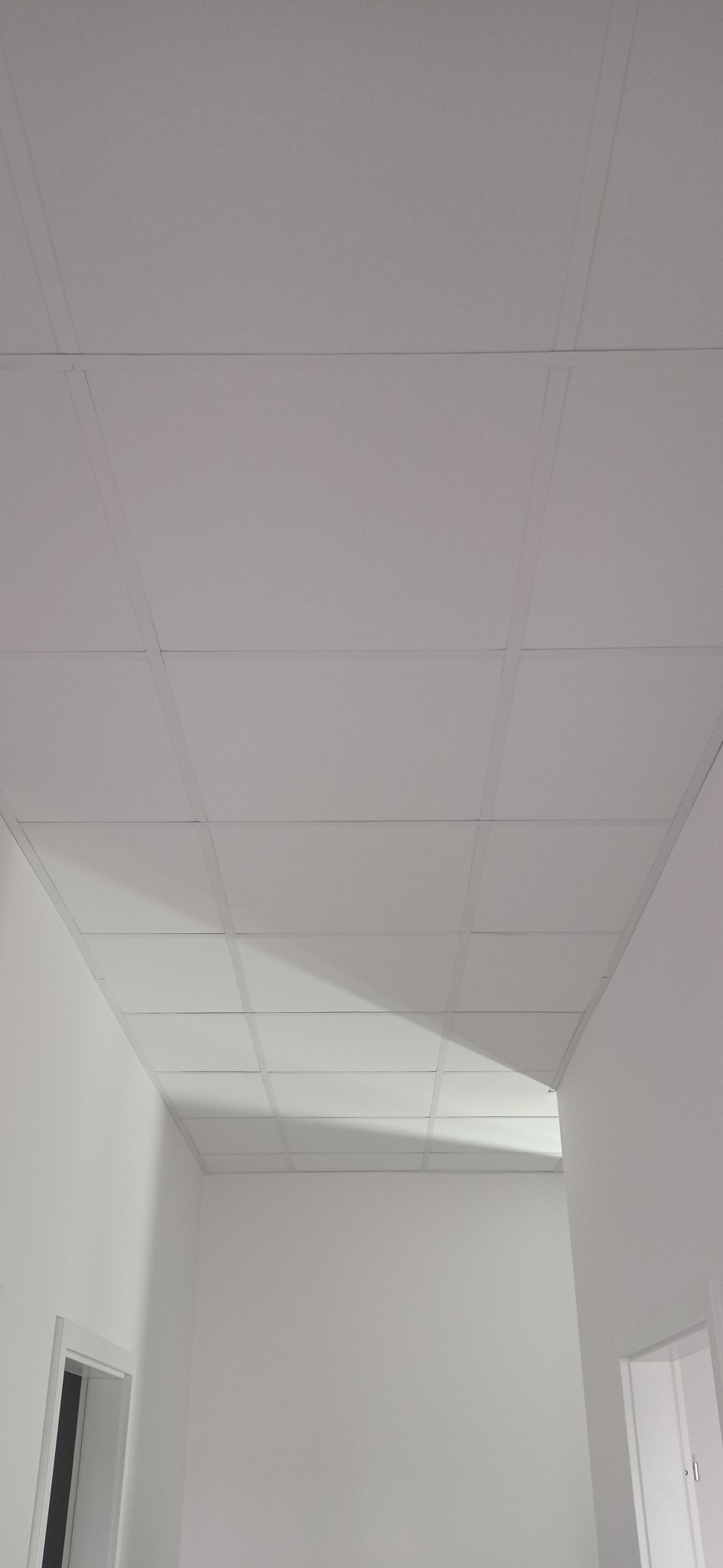 Biały sufit podwieszany kasetonowy w nowym pomieszczeniu, widok z dołu, z fragmentami ścian i ościeżnicami drzwiowymi. Minimalistyczny design.