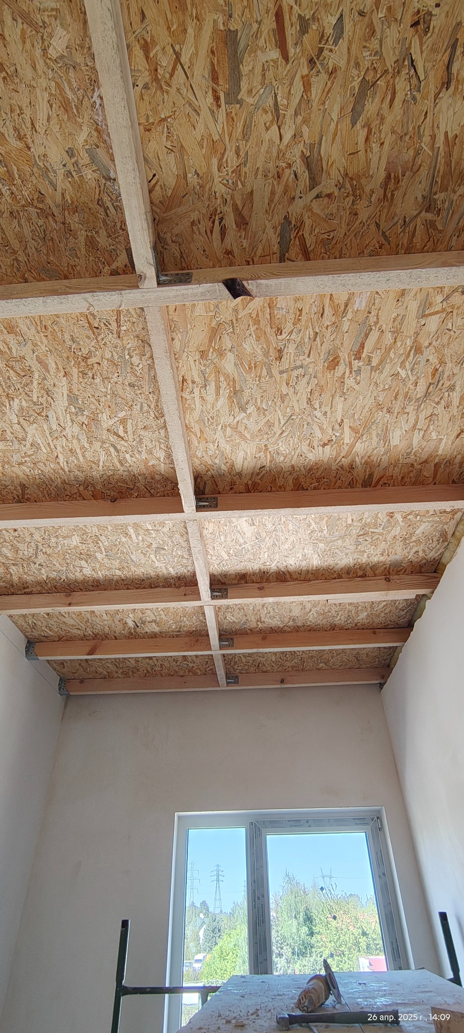 Montaż sufitu z płyt OSB na drewnianej konstrukcji w nowym budynku. Widoczne metalowe łączniki i fragment okna z widokiem na zewnątrz. Surowy stan wykończenia.