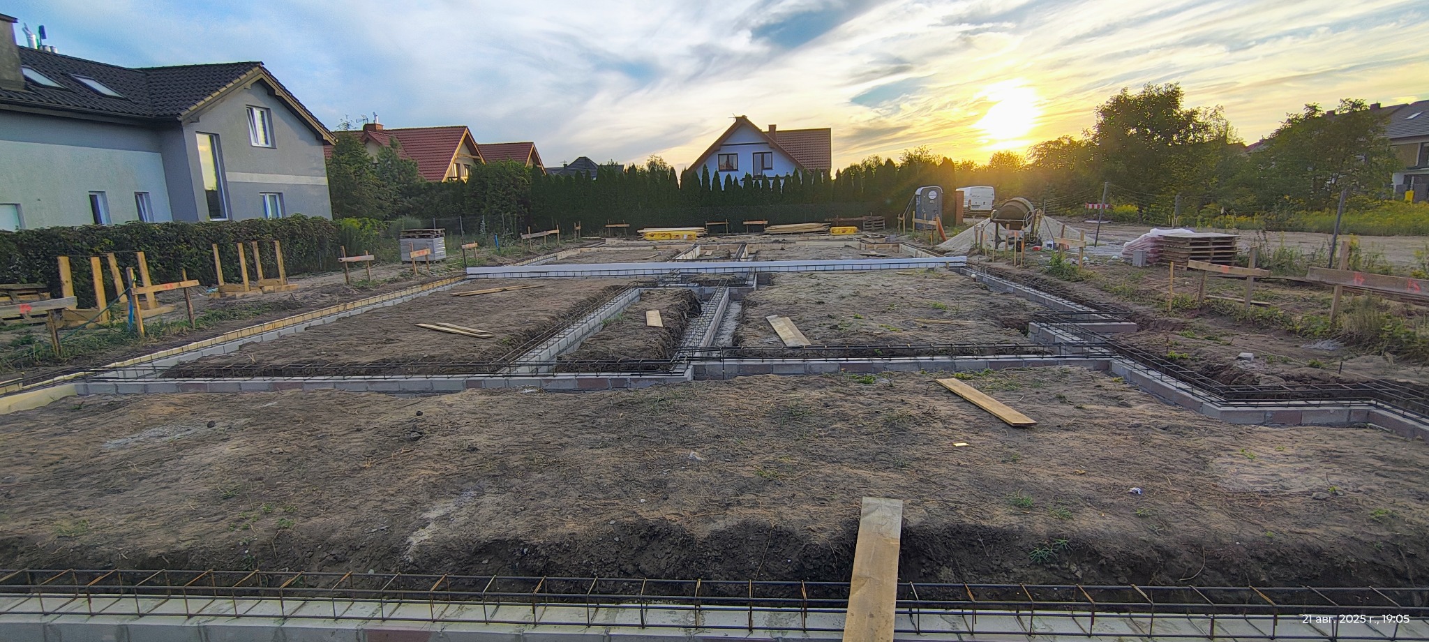 Zbrojenie fundamentów pod budowę domu jednorodzinnego, widok z góry na wykop z ułożonymi prętami zbrojeniowymi i bloczkami betonowymi, w tle zachodzące słońce.