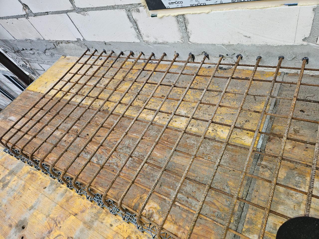 Zbrojenie fundamentu na deskowaniu, widoczne pręty zbrojeniowe i elementy dystansowe. Ściana z bloczków betonowych w tle. Przygotowanie do wylewania betonu.