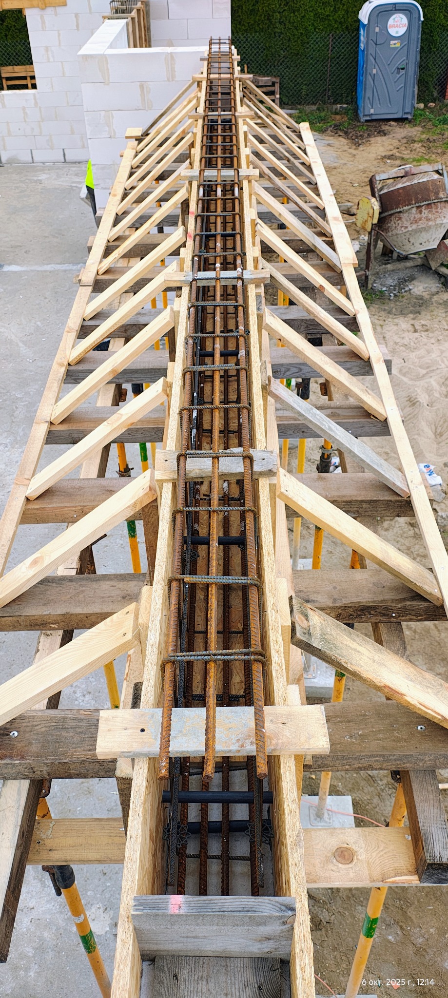 Zbrojenie ławy fundamentowej w drewnianym szalunku, widok z góry. Elementy zbrojenia połączone drutem. W tle bloczki betonowe i przenośna toaleta.