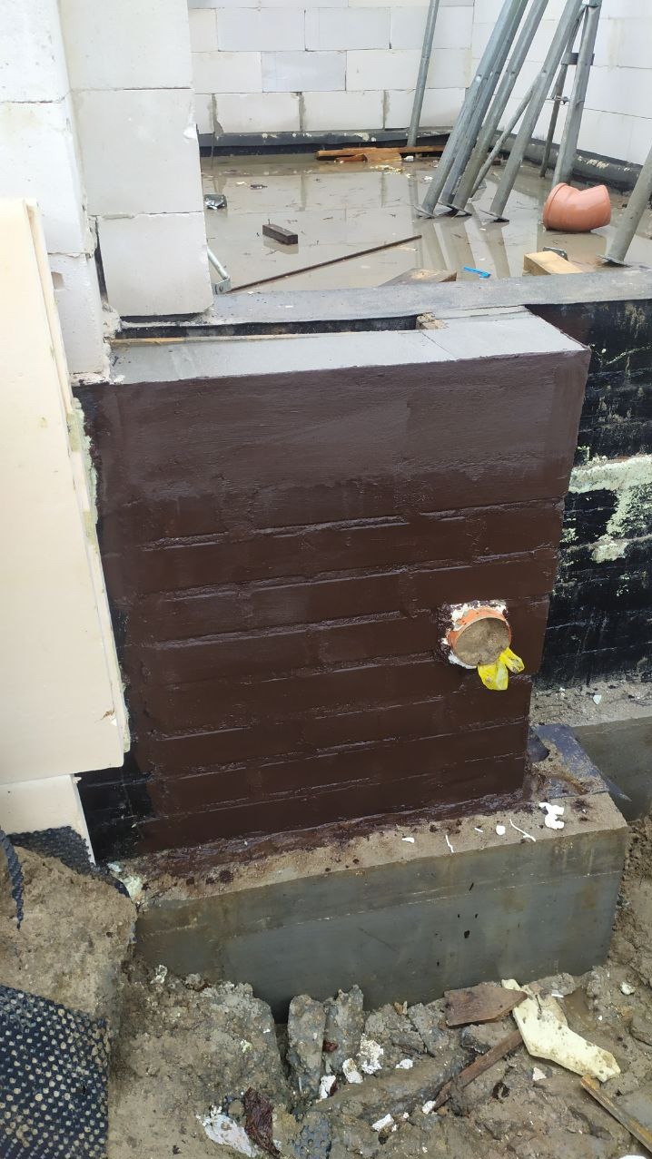 Zabezpieczony, pomalowany na brązowo fragment fundamentu z widoczną rurą odpływową. W tle ściana z białych bloczków i stojące podpory. Prace izolacyjne fundamentów.