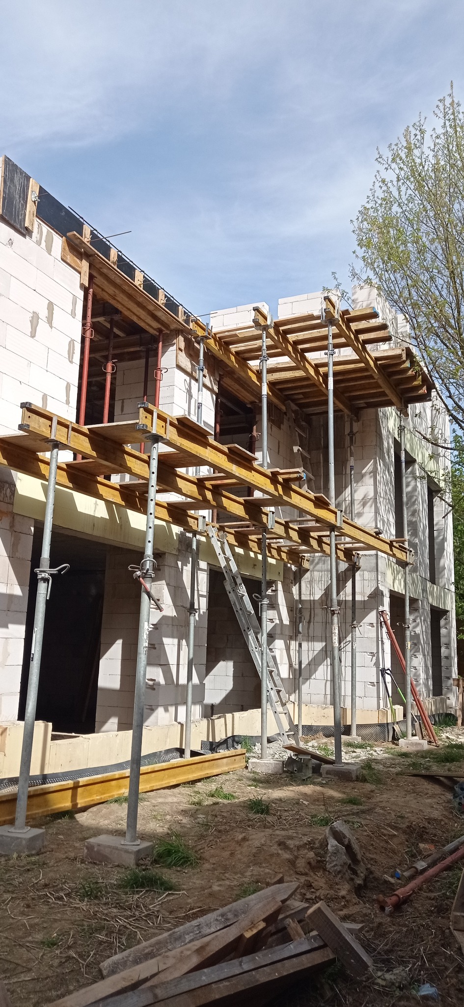 Budowa domu z białych bloczków, widoczne rusztowania i podpory stropowe. Etap murowania ścian i przygotowania do wylania stropu. Ujęcie z dołu, widoczne niebo.