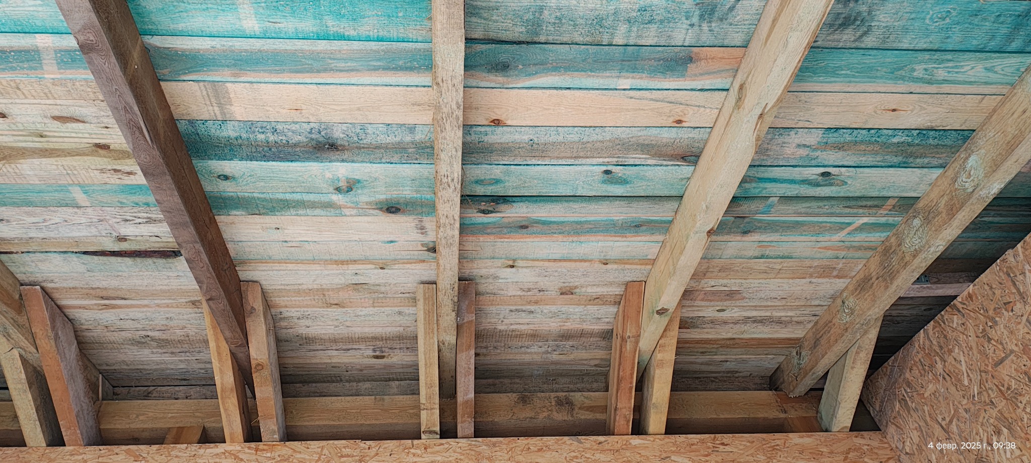 Konstrukcja drewniana dachu z widocznymi belkami i deskami w odcieniach brązu i turkusu. Elementy z płyty OSB w tle. Widok z dołu, surowy stan wykończenia.