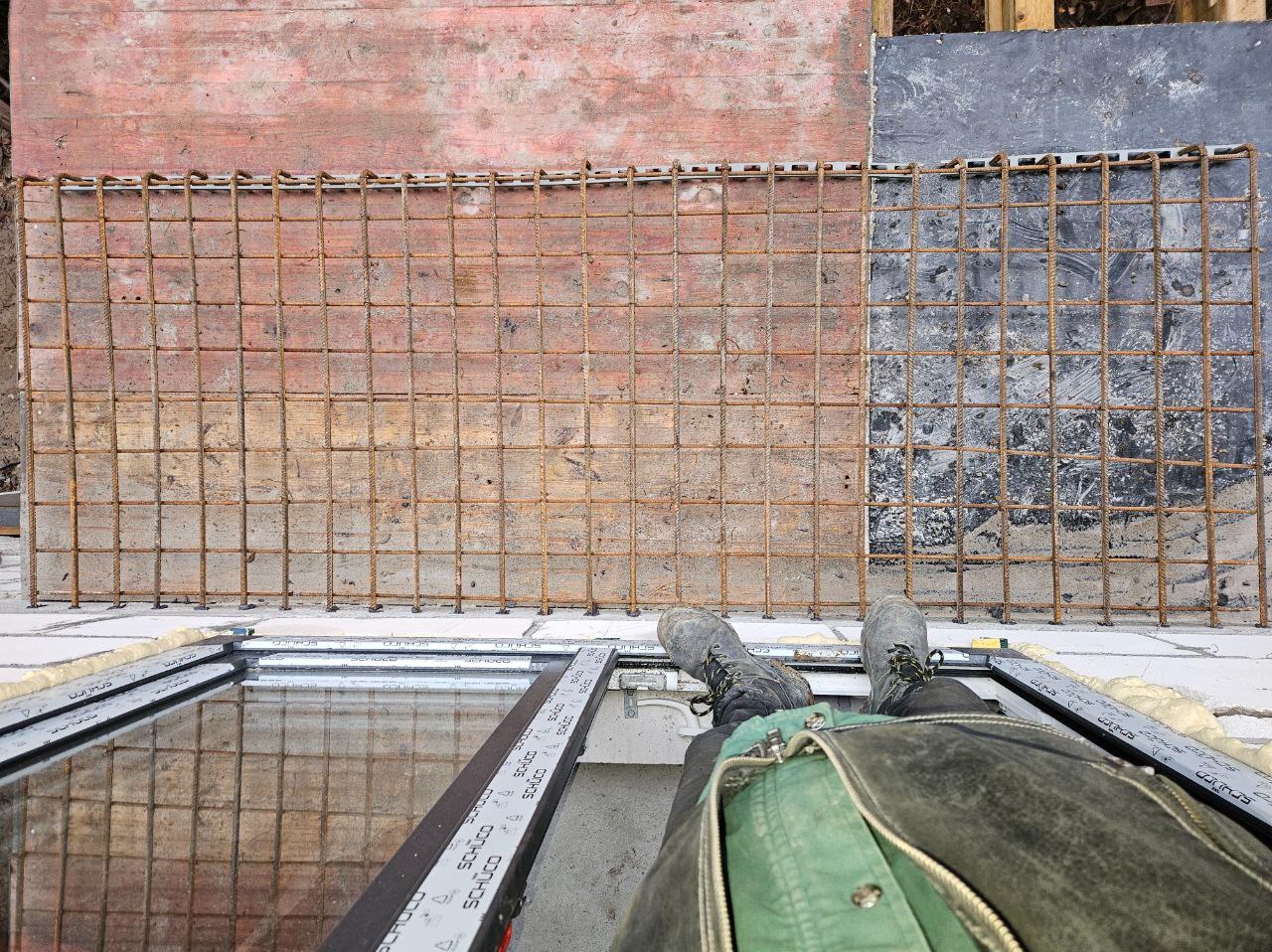 Zbrojenie fundamentu z widocznymi butami pracownika i ramą okienną. Siatka zbrojeniowa ułożona na białych bloczkach, przygotowana do zalania betonem.