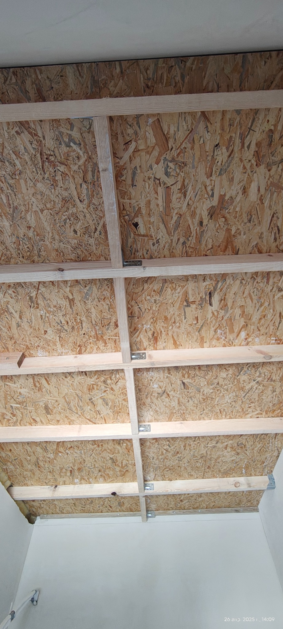 Sufit z płyt OSB i drewnianej konstrukcji nośnej, widok z dołu. Elementy połączone metalowymi łącznikami. Widoczne fragmenty białych ścian.