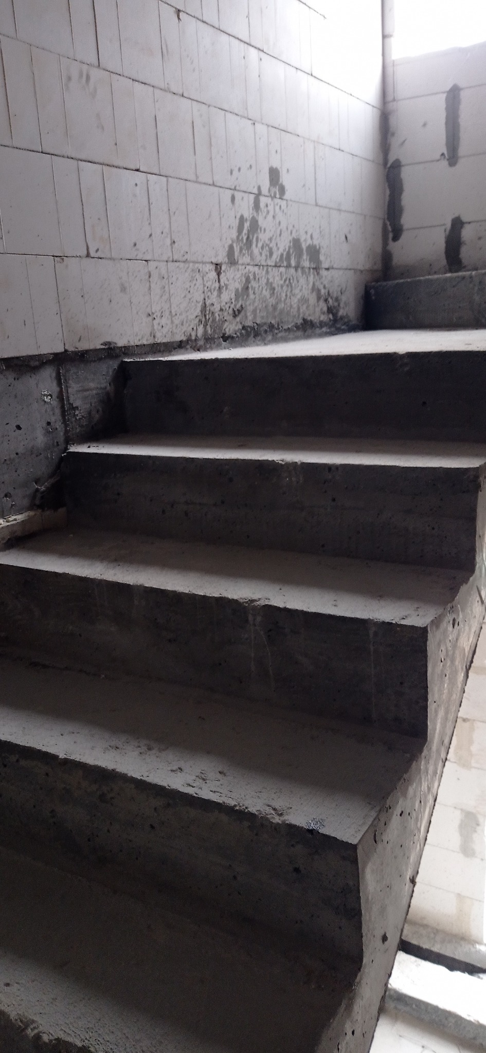 Surowe, betonowe schody w trakcie budowy, z widocznymi blokami betonowymi na ścianach. Ujęcie z dołu, podkreślające surowość i etap prac budowlanych.