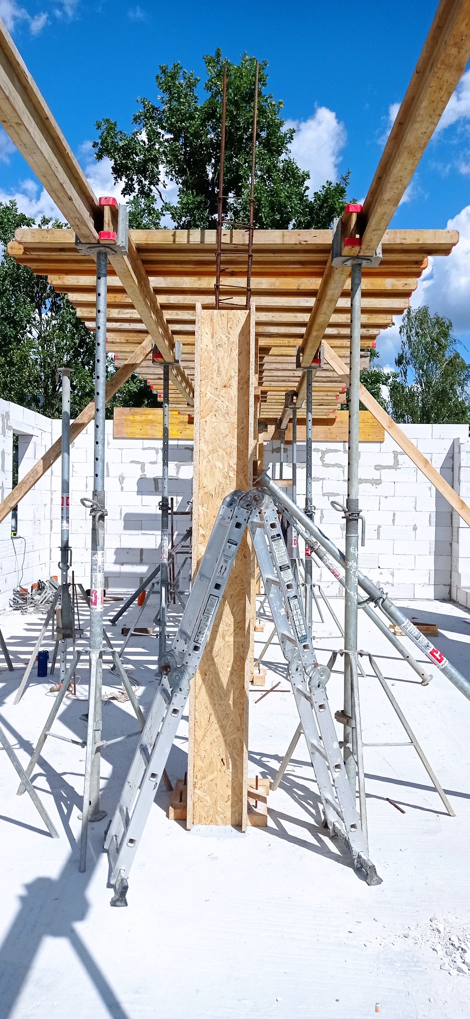 Szczegółowy widok szalunku kolumny na budowie domu z białych bloczków, wspartego na metalowych podporach i drabinie, w słoneczny dzień.