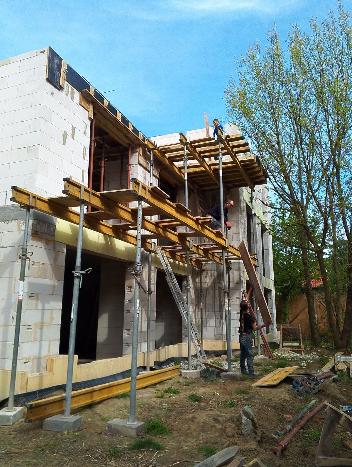 Budowa domu z białych bloczków: widoczne rusztowania, podpory i pracownicy na różnych poziomach konstrukcji. Ujęcie z dołu, błękitne niebo w tle.