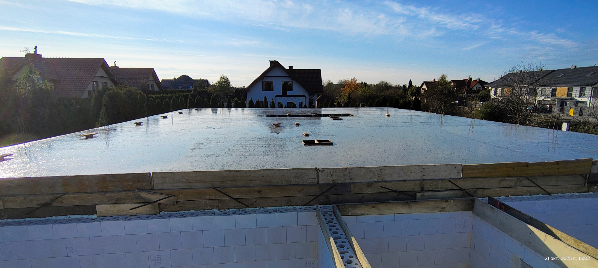 Świeżo wylana, gładka płyta fundamentowa z widocznymi elementami szalunku i zbrojenia na tle zabudowy jednorodzinnej w słoneczny dzień.