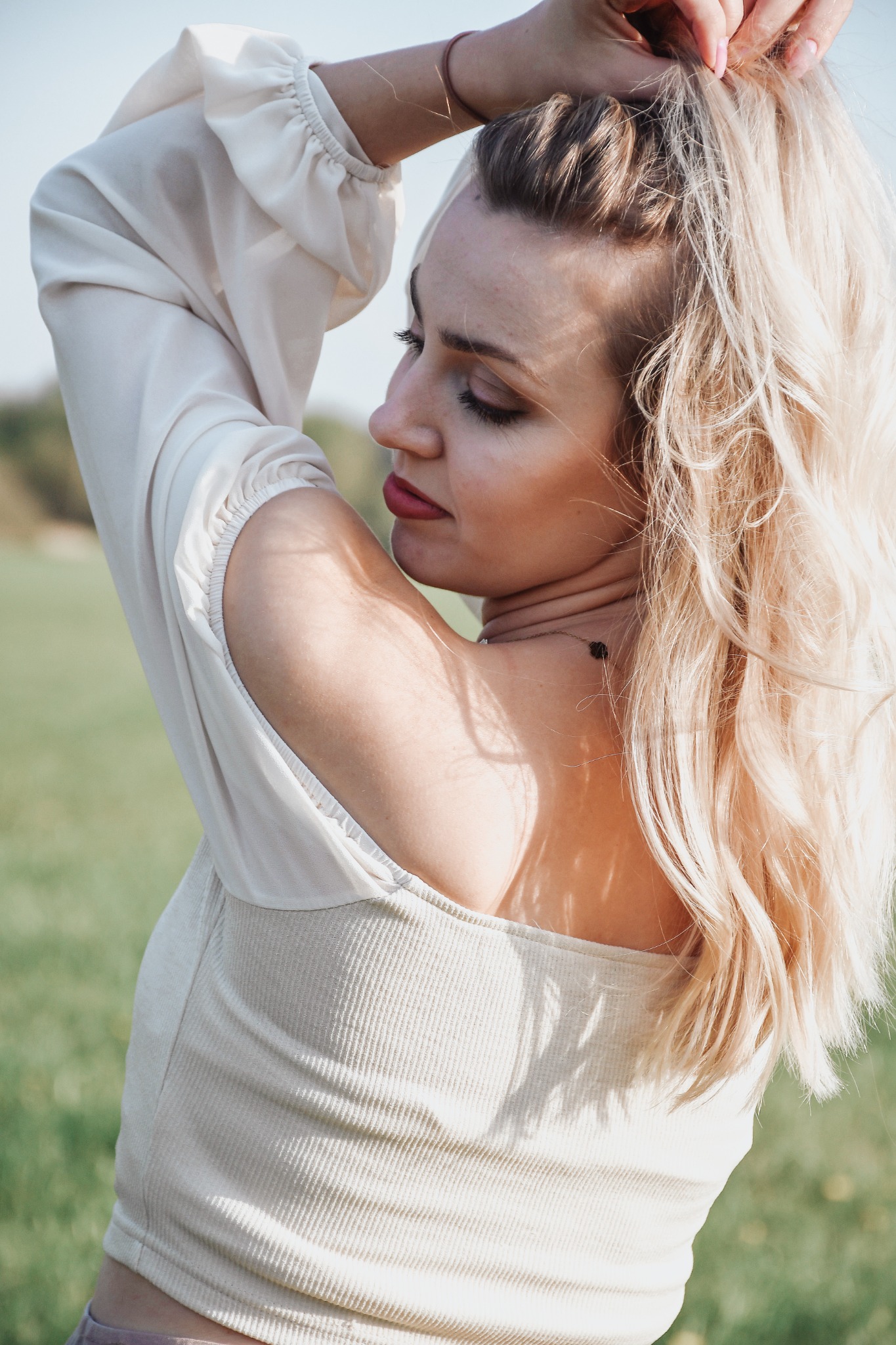 Portret kobiety z blond włosami, ubrana w jasną bluzkę, pozująca na tle zielonej łąki w słoneczny dzień. Widok z profilu, skupienie na twarzy i włosach.