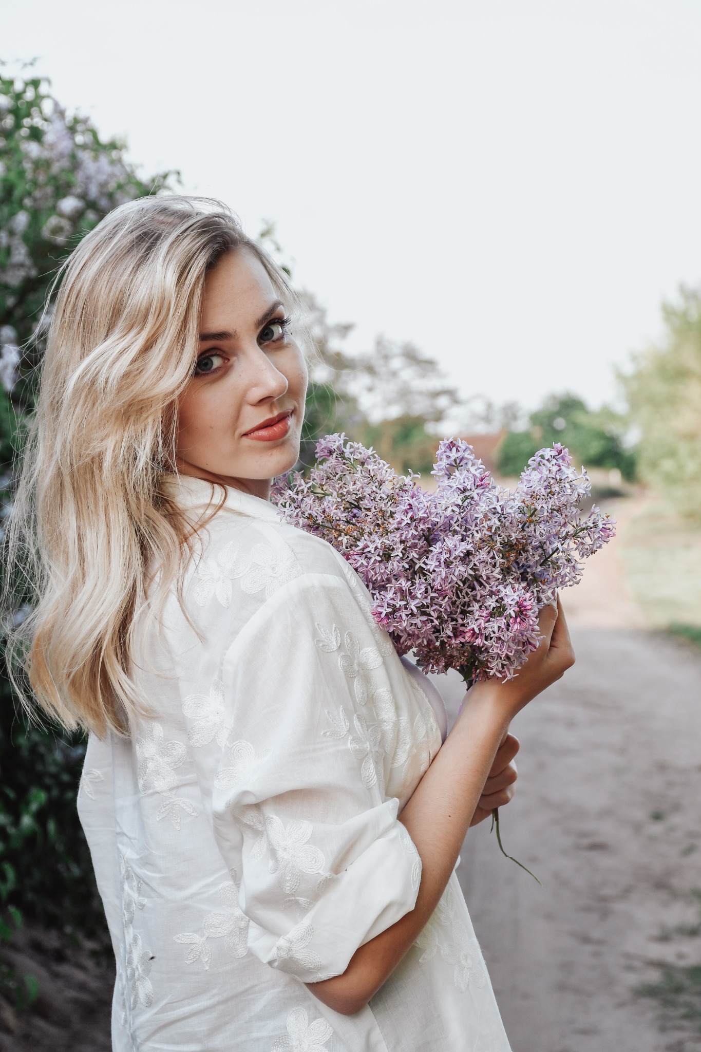 Portret kobiety z blond włosami, trzymającej bukiet bzów na tle wiejskiej drogi w Kcyni. Biała koszula z haftem i delikatny makijaż.