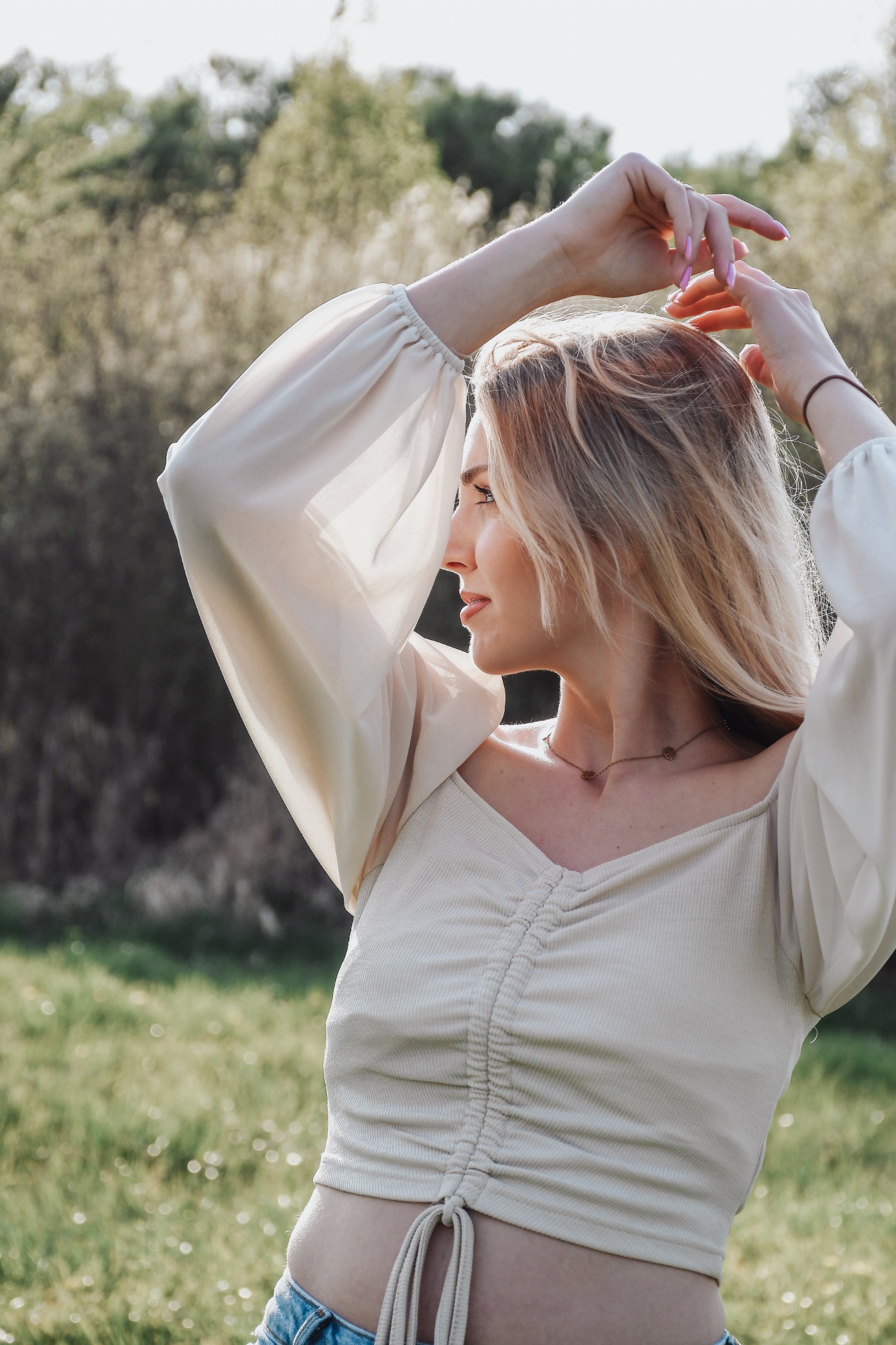 Portret kobiety z blond włosami w jasnej bluzce z bufiastymi rękawami, pozującej na tle zieleni w plenerze. Profil, ręce uniesione do góry.