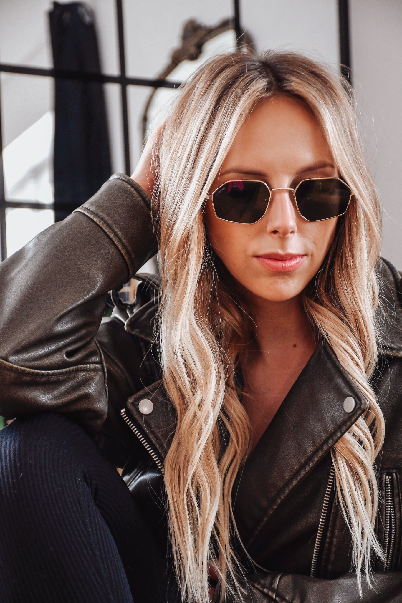 Blond kobieta w okularach i skórzanej kurtce, opierająca głowę na ręce. Ujęcie z bliska, w tle okno z czarnymi szprosami i element dekoracyjny.