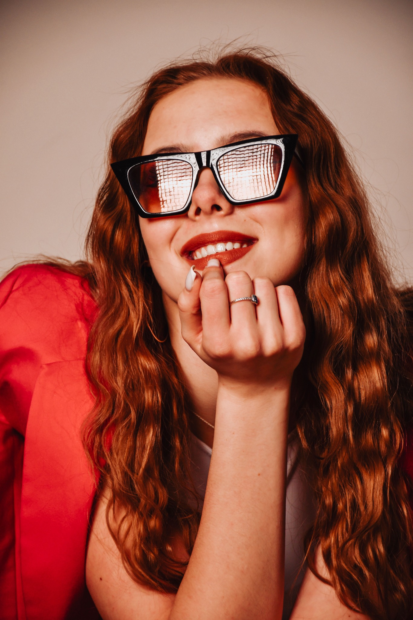 Uśmiechnięta kobieta w okularach cat-eye, czerwonej marynarce i z pierścionkiem, z ręką przy ustach. Widoczne odbicia w szkłach okularów.