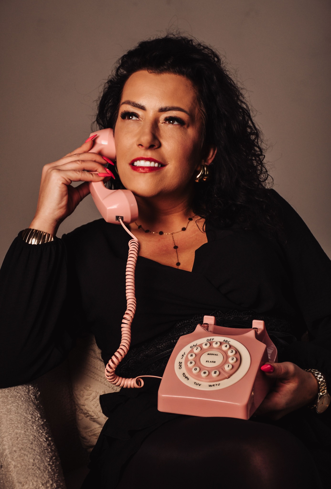 Kobieta w czarnej sukience rozmawia przez różowy telefon retro, spoglądając w górę. Złota biżuteria i czerwone usta dodają elegancji. Stonowane tło.