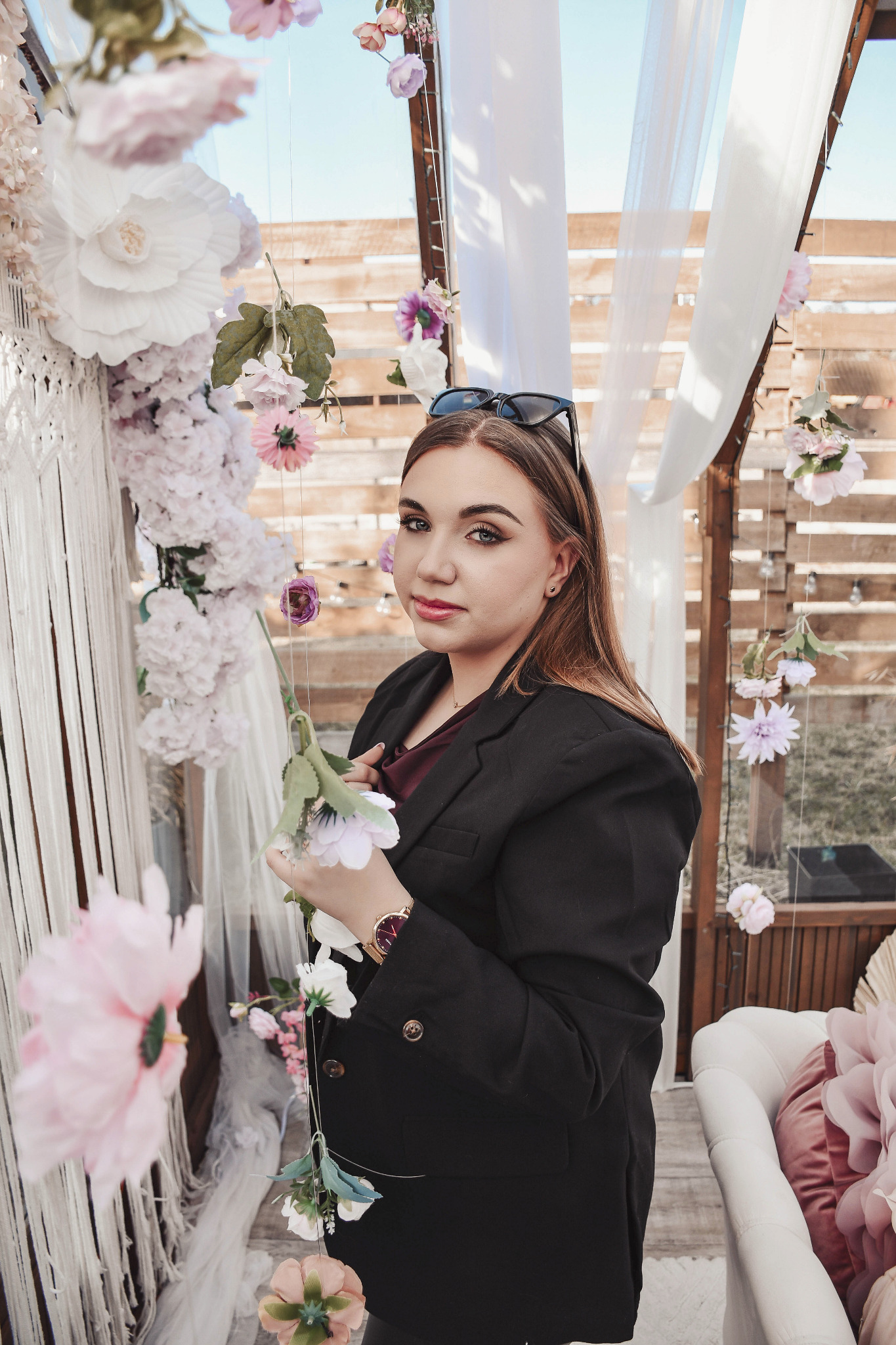 Portret kobiety w czarnej marynarce, trzymającej kwiaty na tle dekoracji kwiatowych i białej tkaniny, z okularami na głowie. Ujęcie z dołu.